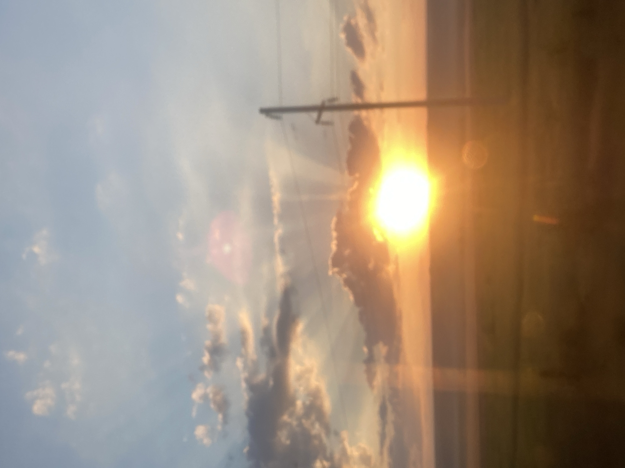 The image depicts a blurry sunset over a field in Balabanu, Moldova. The sun is low on the horizon, casting a warm orange glow across the sky and the land. The sky is filled with scattered clouds, some of which are illuminated by the setting sun. A utility pole stands to the right of the frame, with power lines stretching across the sky. The foreground is a blurred expanse of what appears to be a field. There are no people visible in the image. The overall impression is one of a tranquil, natural landscape at dusk.