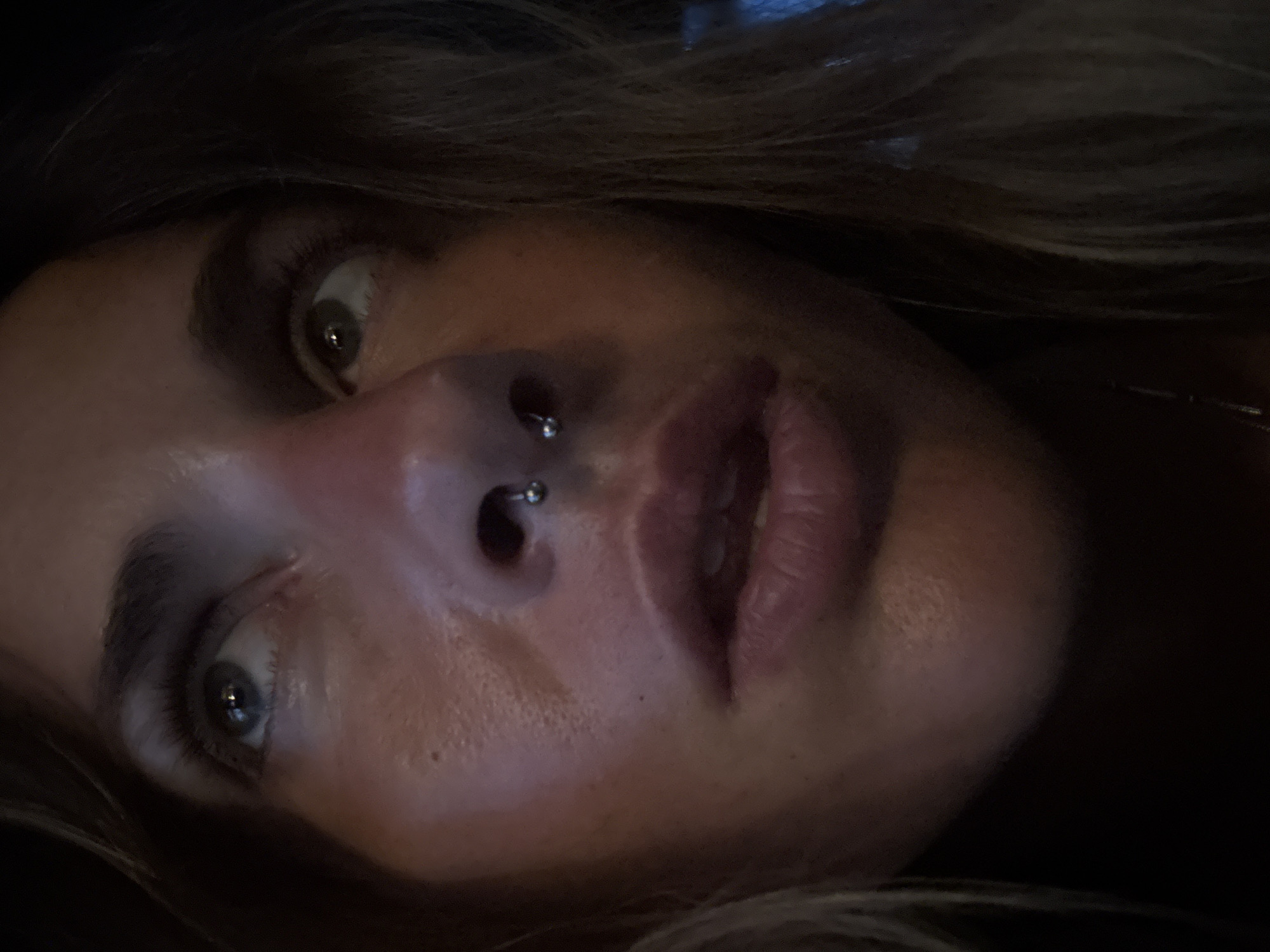 A close-up, low-angle, outdoor shot shows the upper portion of a Caucasian woman's face. Her hair, a light brown with blonde highlights, is partially visible on the left and right sides of the frame, obscuring the background. She is looking upwards and to her left. Her left eye is a light blue-grey, and her right eye appears to be a hazel or green color. She has dark eyebrows and lashes. A silver septum piercing with a small ball on each end is visible in her nose. Her lips are full and painted with a matte, peachy-pink lipstick. Her skin is sun-kissed with visible pores and some subtle blemishes. The lighting suggests it is dusk or night.