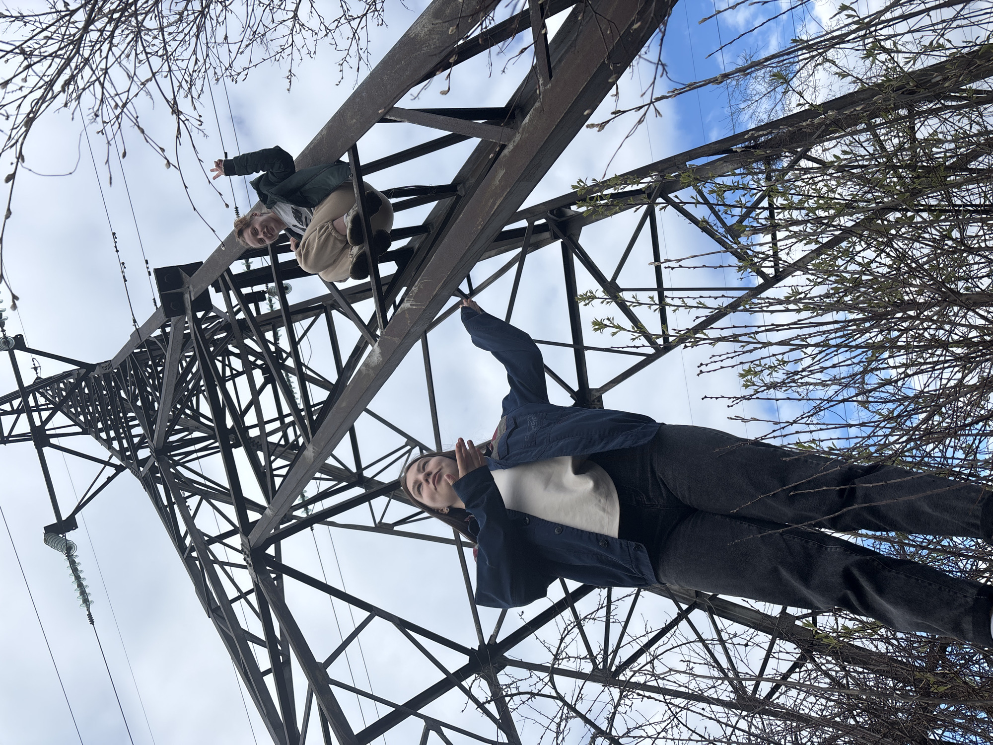 Two people are atop an electrical transmission tower. One is squatting with their left arm raised, and the other is standing, also with their left arm raised. The photograph is taken from below, looking up at the tower and the people. The sky is visible between the branches of trees and the tower structure, and the tower is constructed of metal. This image was taken near Chaikovsky, Russia.