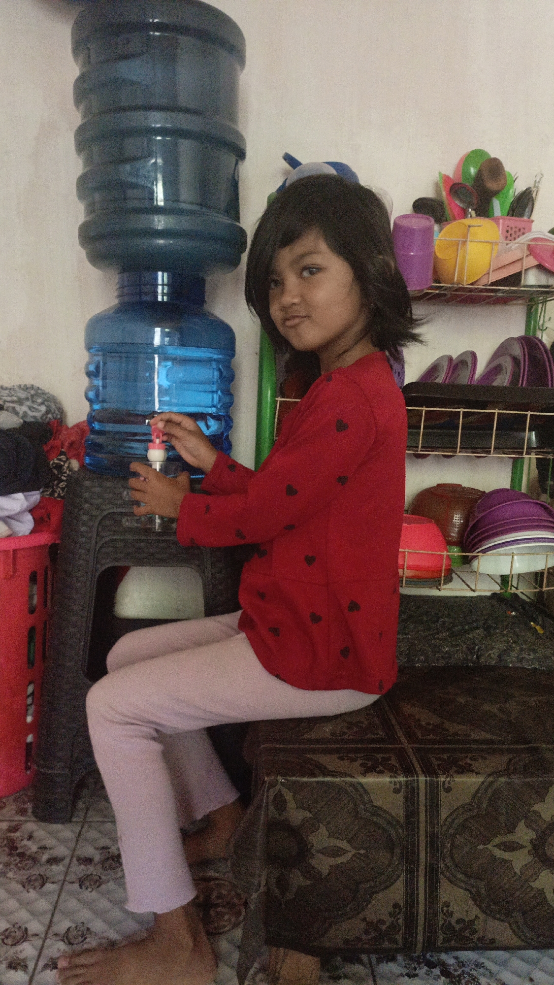 A young girl with dark hair and eyes is sitting on a patterned bench next to a large blue water cooler in a room. She is wearing a red long-sleeved shirt with small black hearts and light pink pants. Her right hand is reaching towards the water cooler dispenser. Behind her, a shelf holds various items, including colorful containers and what appear to be plates or bowls. A red plastic bin is visible to her left. The floor is tiled with a floral pattern. The wall behind the water cooler is a light color. The scene appears to be indoors, possibly in a home or a communal area in Pejala, Indonesia.