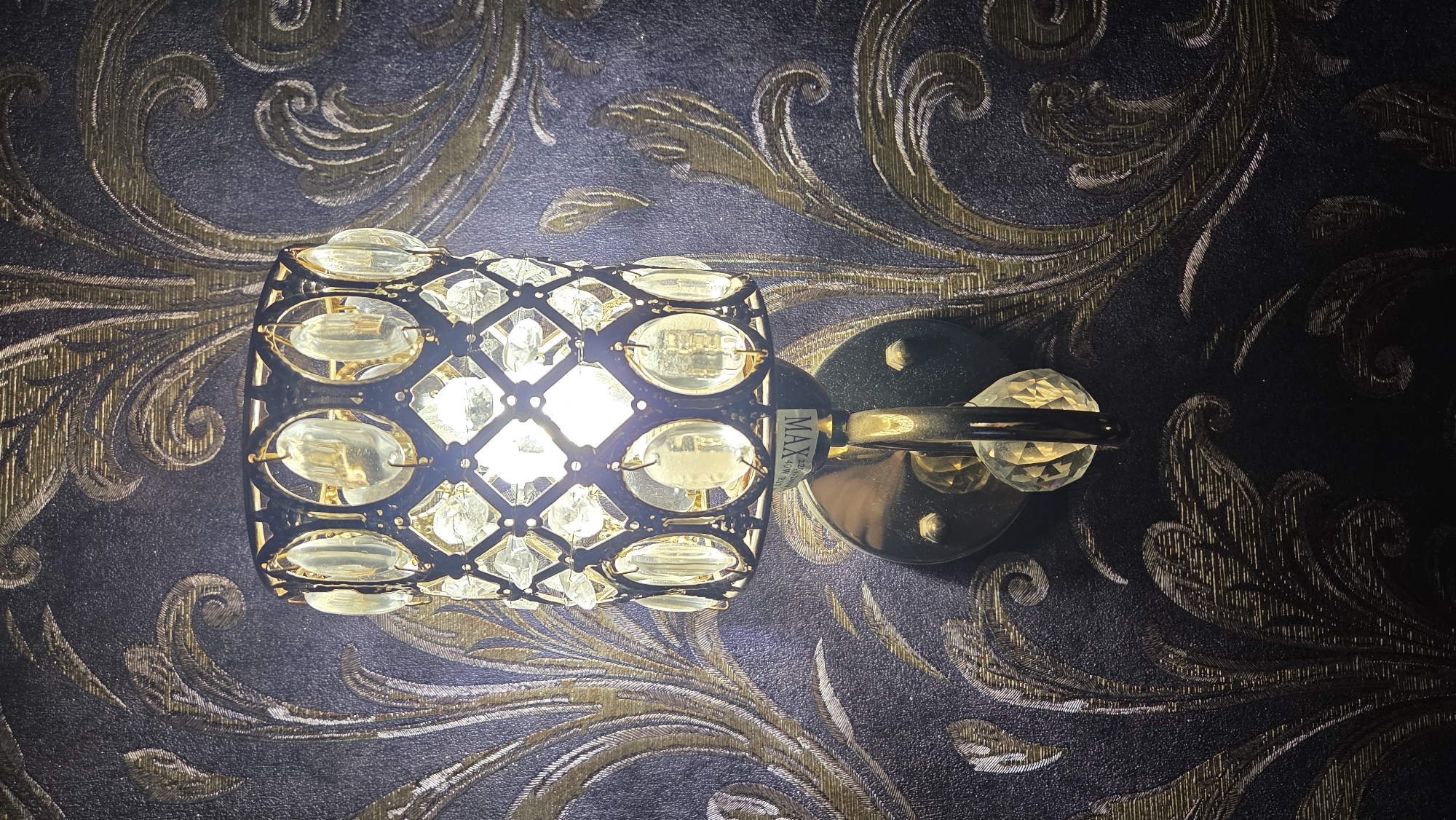 A close-up, vertical shot depicts an illuminated decorative wall sconce mounted on a textured, ornate wallpaper in an interior setting in Chaikovsky, Russia. The light fixture features a dark-colored metal frame forming a cage around a bright, central bulb. This frame is adorned with numerous clear or slightly amber-tinted faceted crystals or glass pieces, arranged in a grid and oval patterns, which refract the light. The light source itself glows brightly, casting a warm hue.
The sconce's base consists of a round, metallic gold-toned plate attached to the wall, with a small, faceted crystal sphere extending from it, enhancing the fixture's opulent design.
The wallpaper behind the sconce has a rich, dark background, possibly deep grey or black, embellished with elaborate, swirling patterns in a metallic gold or bronze finish. These intricate designs resemble paisley or damask motifs, giving the wall a luxurious and classic appearance. The light from the sconce illuminates the immediate area, accentuating the texture and sheen of the wallpaper and creating subtle shadows.
A small label is visible on the light fixture's base, displaying text that includes "MAX," "220-240V," and "40W E14," indicating its maximum wattage, voltage, and bulb type. The artificial illumination suggests the scene is set in the evening or night, or within a dimly lit room, contributing to an atmosphere of elegance and warmth.