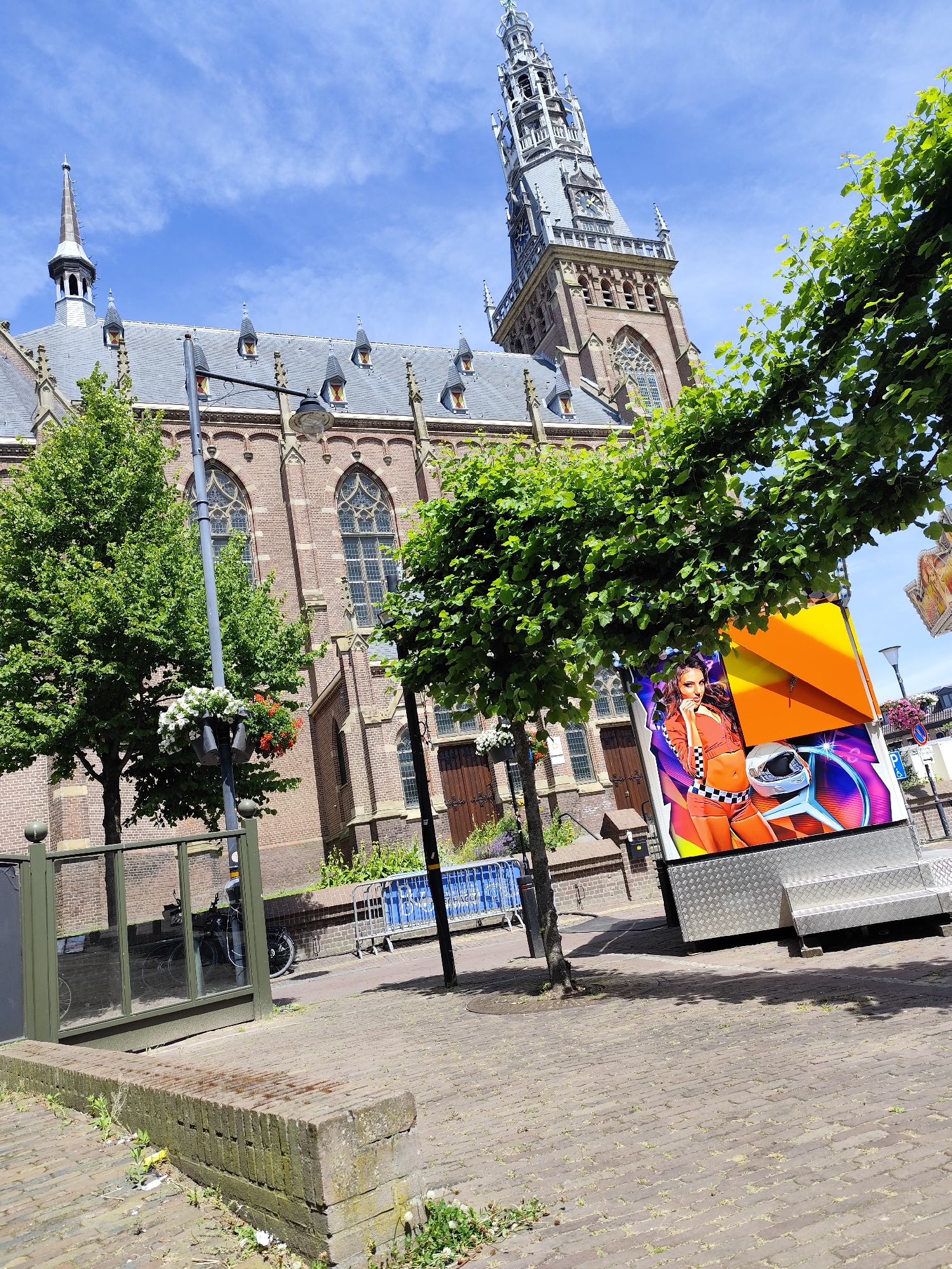 The image shows a view of a town square in Schagen, Netherlands, with a large brick building featuring a prominent clock tower. In the foreground, a colorful, modern trailer with an image of a woman in racing attire is parked on a brick-paved area. Trees with green foliage are scattered throughout the scene, casting shadows on the ground. A lamppost with hanging flower baskets stands near the trailer. In the background, a portion of the historic building, likely a church or municipal building, is visible, characterized by its brick facade and Gothic-style windows. A blue barrier fence is present in the middle ground. The sky is blue with scattered white clouds.