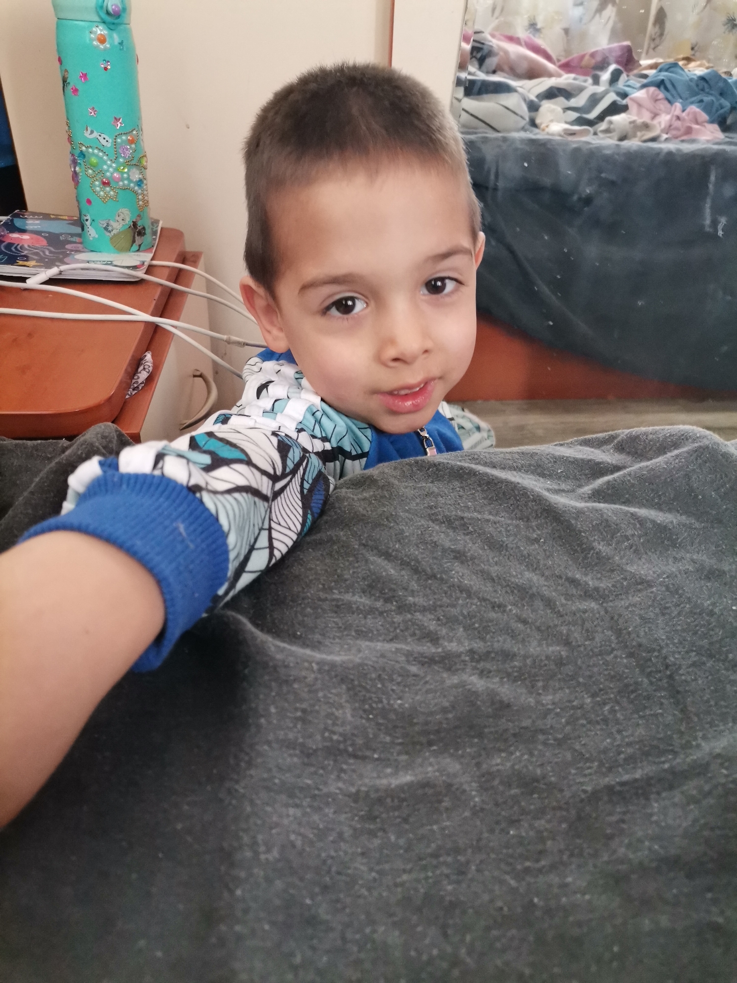The image shows a young child with short brown hair and brown eyes looking at the camera. The child is wearing a blue and white patterned shirt with a blue cuff. The child appears to be resting on a gray blanket. Behind the child is a light brown side table with various items on it, including a turquoise water bottle decorated with stickers, papers, and white cords. In the background, there is a bed with assorted clothing and blankets. The location is unknown.