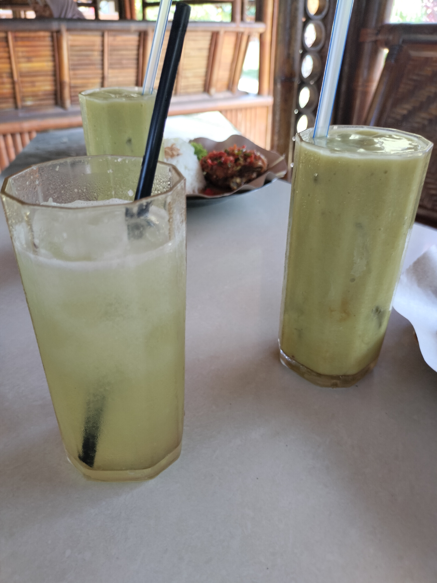 Three drinks are displayed on a light-colored table. The drink in the foreground is in a clear, faceted glass and appears to be a light green, frothy beverage with ice and a black straw. Behind it, a similar glass contains a pale yellow, frothy drink with ice and a straw. To the right, a taller, clear glass holds a thick, pale green liquid with ice and a blue and white striped straw. A small plate with rice and what appears to be fried food topped with chili sauce is visible in the background. The setting is an open-air structure with bamboo walls and roof.