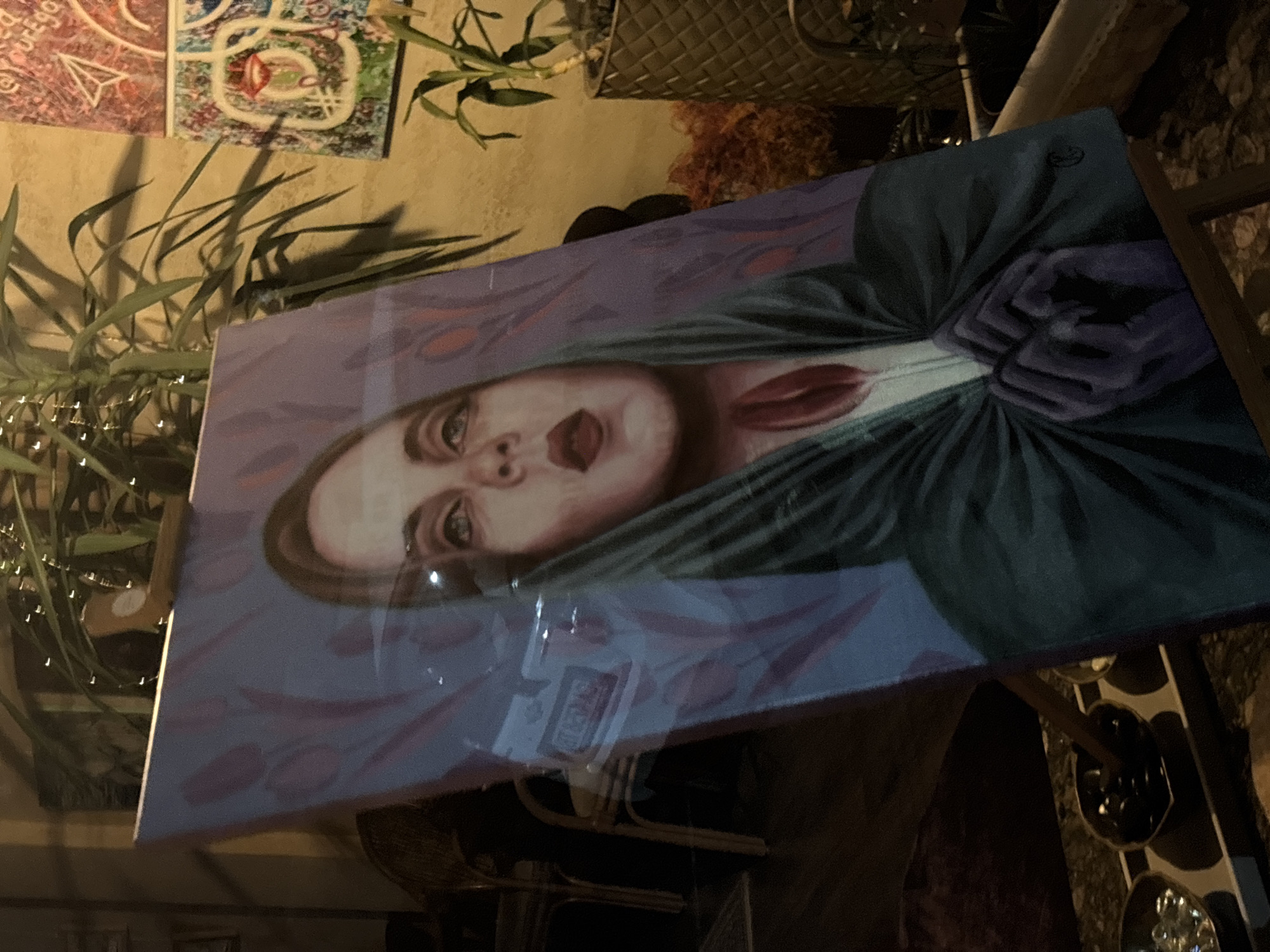 The image shows a painting displayed indoors, likely in an art gallery or a private collection. The painting depicts a woman with fair skin, red lips, and dark hair, partially covered by a dark green cloth or garment. The background of the painting is a light purple color with a subtle pattern of flower shapes. The painting is propped on a stand, possibly made of wood or metal. The stand is positioned near a wall that has other artwork and plants decorating the space. The setting appears to be a dimly lit interior. The presence of a lightbulb garland suggests the interior could be a hip, modern cafe. The location is Sofia, Bulgaria.