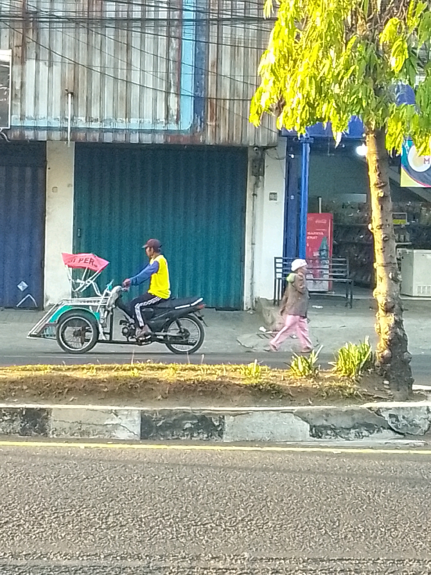 A man wearing a yellow vest and a dark red cap rides a black motorcycle with a sidecar. The sidecar is green with decorative trim and has a pink tarp attached to the front. The man is on a paved road, with a grassy area and a sidewalk to his right. Behind the sidewalk is a building with corrugated metal siding and a dark blue roll-up door. Further down the street, to the right of the building, is a tree with bright green leaves and a person in pink pants walking away from the camera. This scene is located on Jalan Gajah Mada in Jember, Indonesia, likely in front of shops or businesses.