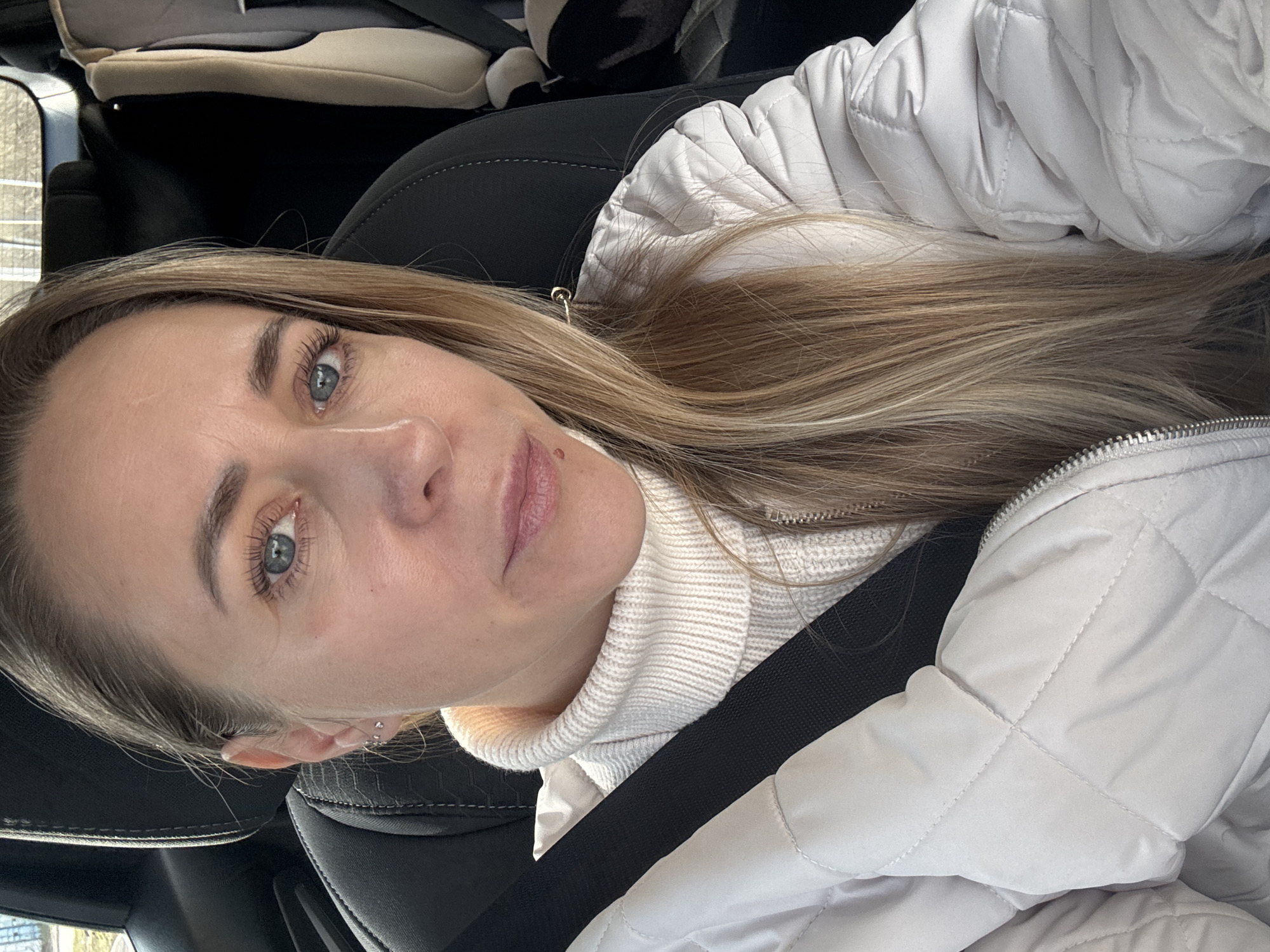 A fair-skinned woman with blonde hair is in the front seat of a car, wearing a white turtleneck sweater and a white quilted jacket, and a black seatbelt across her chest. Her face is angled slightly to the left, and she is looking directly at the camera. The interior of the car includes black seats and a child seat in the back. The location in Chaikovsky, Russia cannot be determined from the visual information.