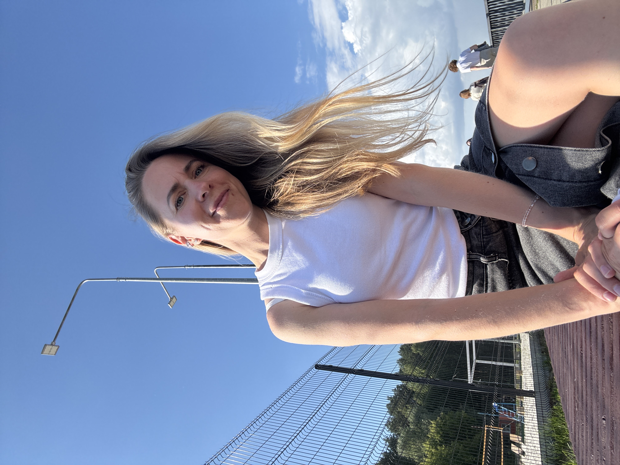 A medium-shot selfie shows a Caucasian woman with long blonde hair and blue eyes, wearing a white tank top and a dark denim skirt, sitting on a wooden bench. She is smiling and looking at the camera, with her right arm resting on her lap. The background features a clear blue sky with a few white clouds, a tall lamp post, a chain-link fence, and some trees. In the distance, two other people are walking on a boardwalk. The scene appears to be an outdoor recreational area, possibly near a body of water, given the presence of a boardwalk and open sky. The woman's attire suggests a casual, warm-weather setting. The image was taken in the city of Chaikovsky, Russia.