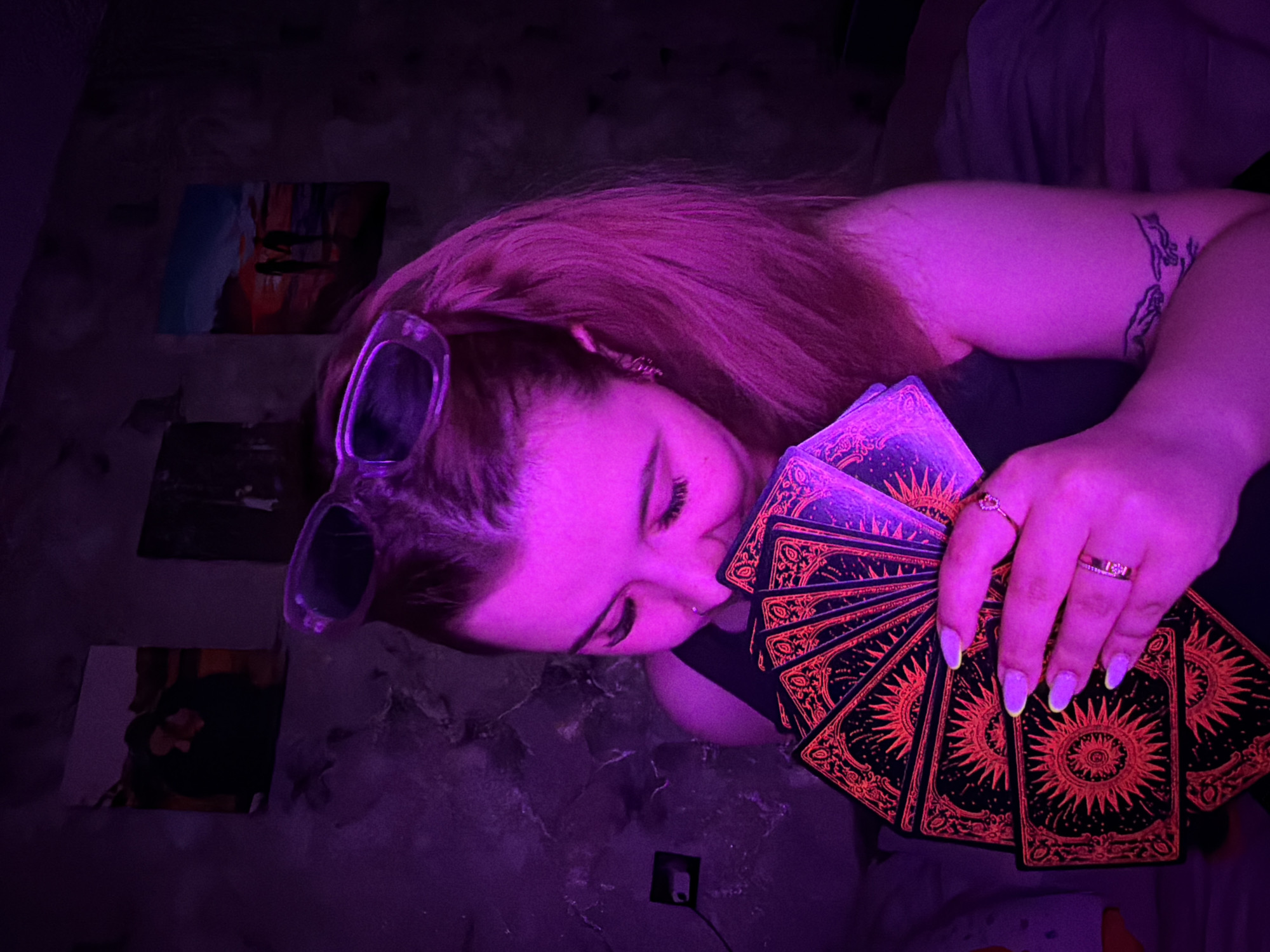 A young woman is lying down, possibly in Chaikovsky, Russia, considering the prompt. She is holding a deck of tarot cards with orange sun-like designs on the backs in her left hand, which has rings on several fingers. Sunglasses are perched on her head. There are several pictures or paintings on the floor/surface next to her. A tattoo is visible on her arm. The scene is lit with a purple light.