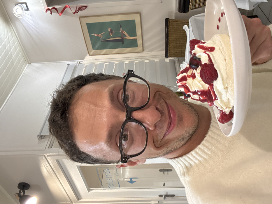 A man with dark hair and glasses smiles, holding a white plate topped with a slice of cake. The cake is adorned with white cream, fresh raspberries, blueberries, and a red sauce. The man is wearing a cream-colored turtleneck sweater. Behind him, a door is visible with the word "TOILETTES" written on it. A framed painting hangs on the wall, and a small fan is attached to the ceiling. A woven chair and white curtain are also visible in the background.