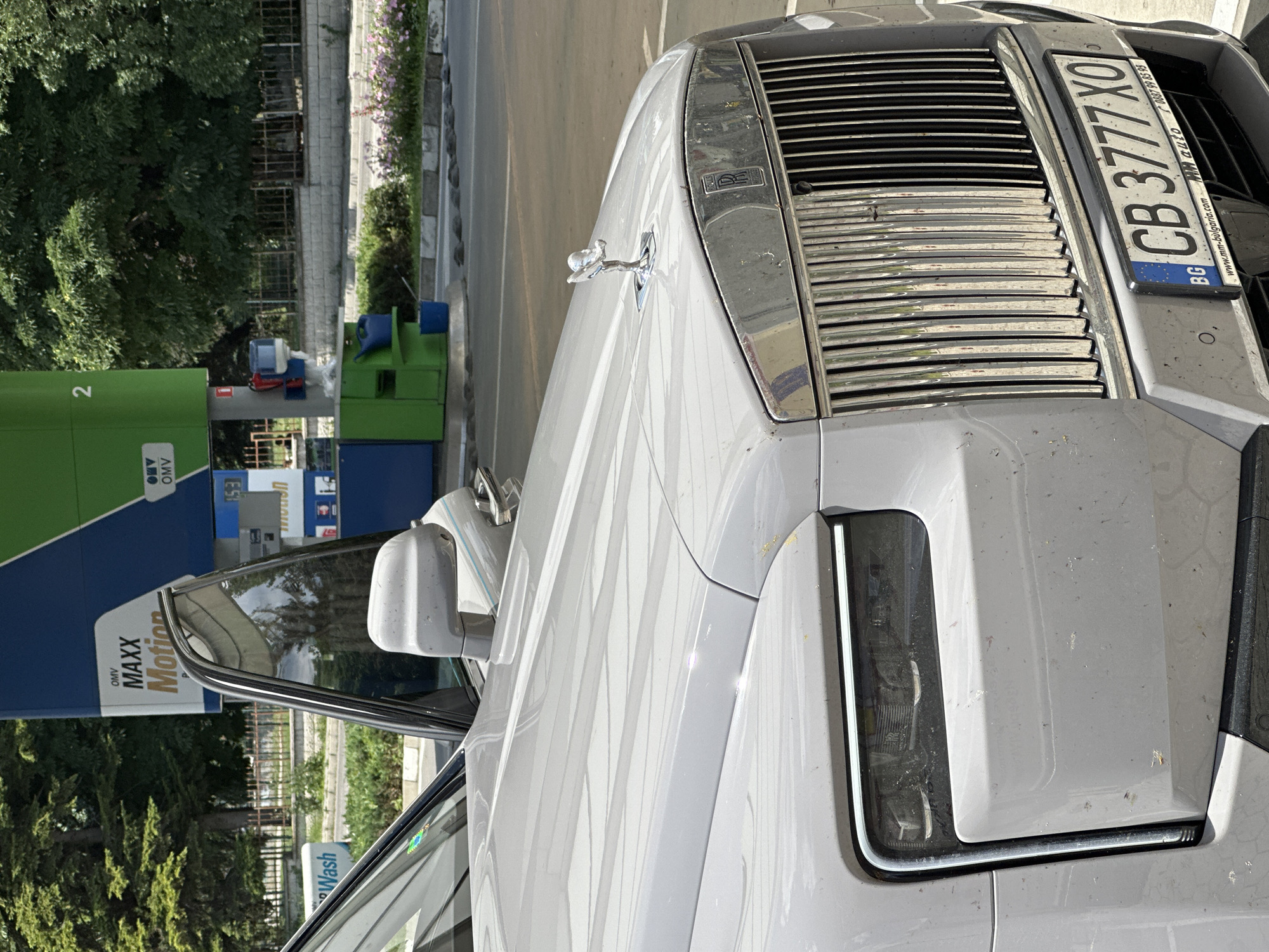 The image captures a close-up view of a light gray Rolls-Royce car parked in front of an OMV gas station. The gas station features blue and green signage with text such as "OMV MAXX Motion" and numbers "2" and "1". The car's distinctive hood ornament, the Spirit of Ecstasy, is prominently visible. The car has a silver grille and a license plate that reads "CB 3777 XO" with "BG" indicating Bulgaria. The background includes trees, a fence, and some flowering plants. The overall scene suggests the car is either refueling or has just completed refueling. The specific location is likely Veliko Tarnovo, Bulgaria, as indicated by the license plate and common gas station chains in the region.