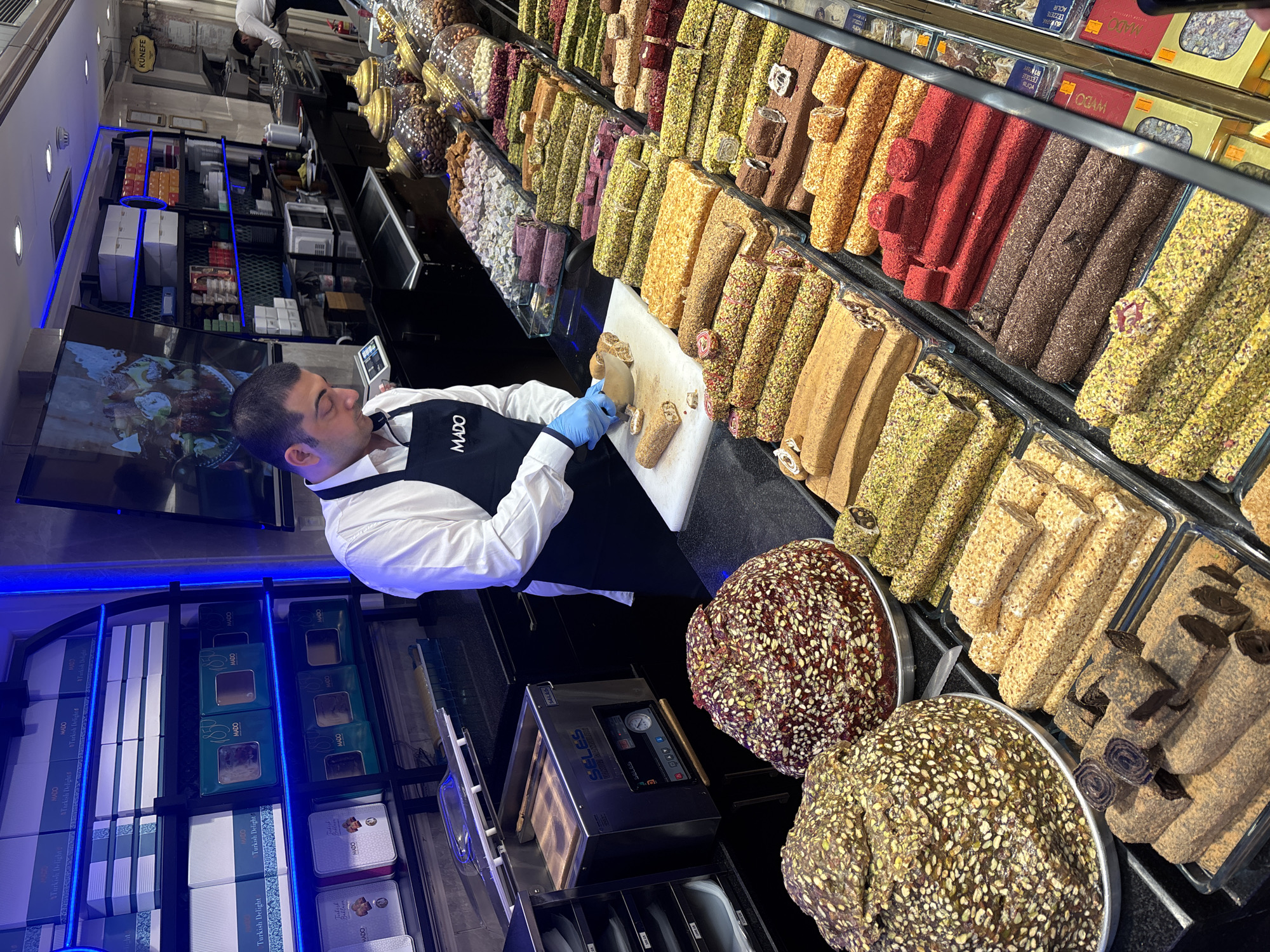 This media file captures a man, wearing a white shirt and a dark apron with the logo "MADO" printed on it, actively preparing Turkish Delight in a brightly lit store. He is using a knife to slice a piece of the confectionary, wearing blue gloves for hygiene. The store's shelves are filled with an abundant variety of Turkish Delight, showcasing different flavors and textures, including those coated in nuts and pistachios, as well as chocolate-covered varieties. In the background, more products are visible, including packaged Turkish Delight boxes. The lighting is artificial and appears to be from overhead fixtures and blue LED strips along some shelves. The overall scene suggests a bustling retail environment, likely a confectionary shop, possibly in Beyoğlu, Turkey, given the "KÜNEFE" sign and the "MADO" branding, a well-known Turkish brand. The man's focused expression indicates he is engaged in his task. There are no indications of the time of day or weather conditions. The presence of a vacuum sealing machine, labeled "SELES," suggests that some products may be packaged for sale or freshness.
