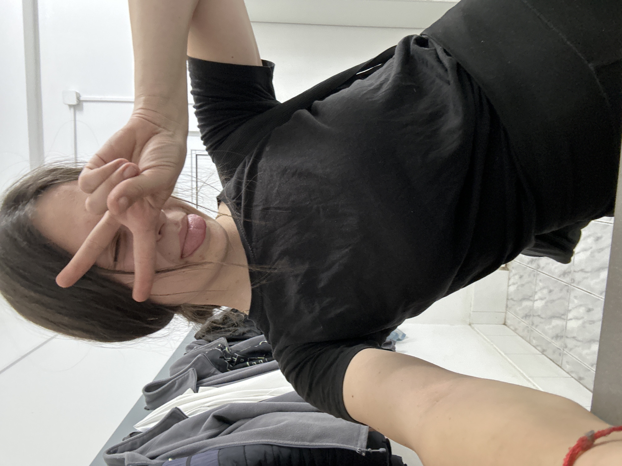 A woman with dark hair is holding up a peace sign while sticking her tongue out. She is wearing a black shirt and pants. The location is uncertain, but the photo may have been taken indoors, near a bed, in Resen, Bulgaria.