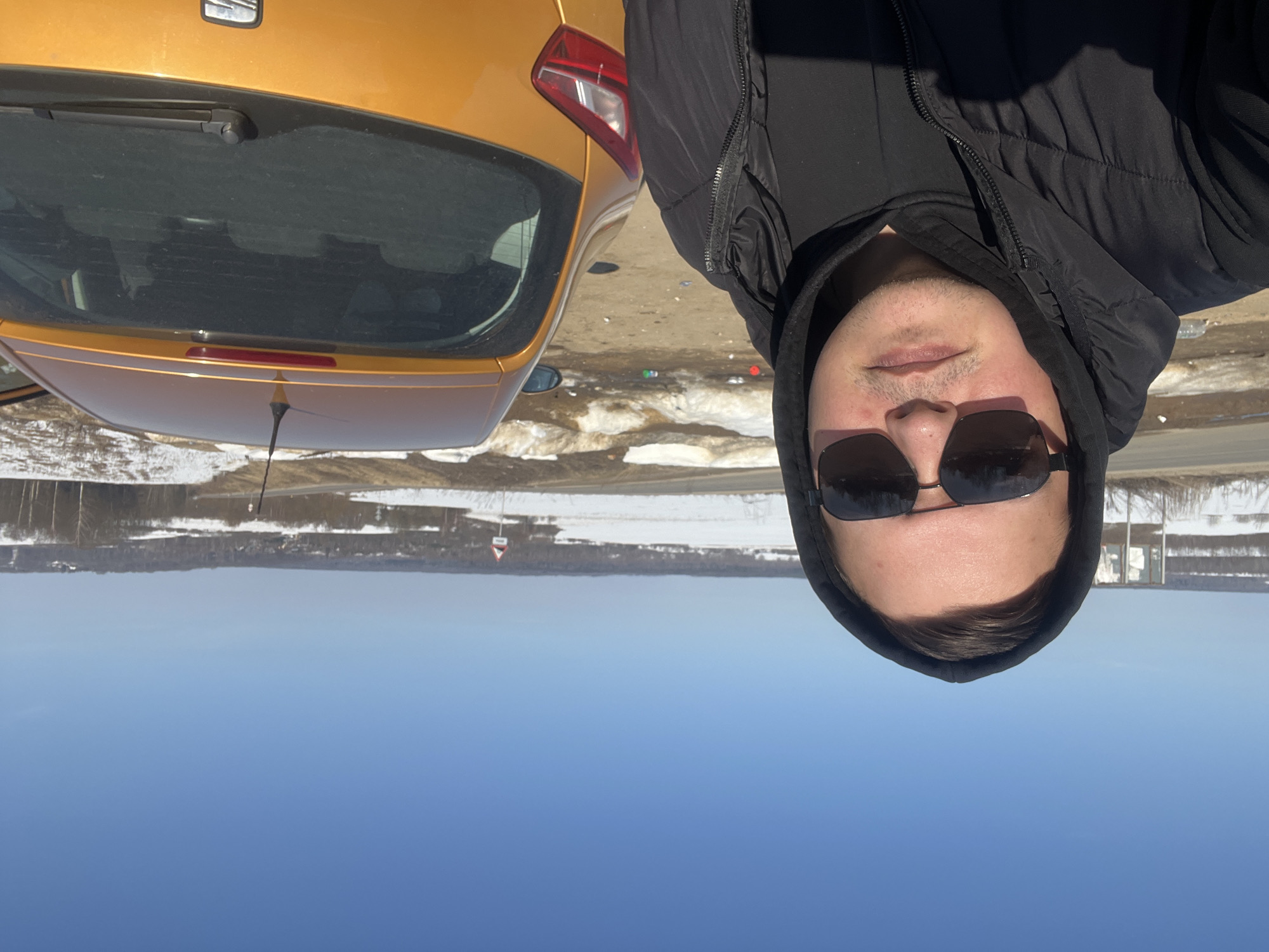 The image captures a male individual positioned in the foreground for a close-up selfie in Zubovo, Russia. The man wears dark sunglasses, a black hooded sweatshirt with the hood up, and a black puffer vest, looking directly at the camera. To his right, the rear of a gold/orange hatchback vehicle is visible, parked on an unpaved surface. The vehicle's rear passenger door appears ajar. The immediate ground consists of dirt, mud, and patches of melting snow, with scattered litter visible. A paved road runs diagonally across the midground. Beyond the road, the landscape features snow-covered fields, sparse trees, and distant forested areas under a clear, bright blue sky. A small settlement and a triangular red-bordered warning sign are discernible on the distant horizon.