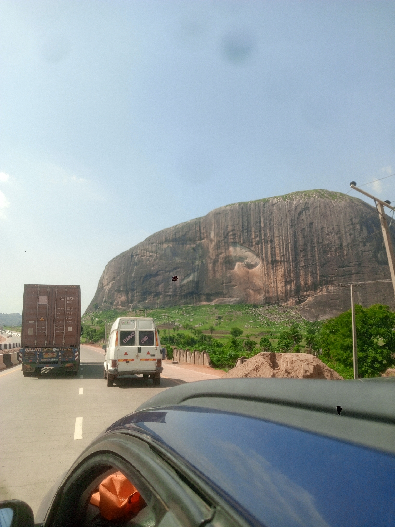 The image shows a view from inside a car driving on a highway. A brown shipping container truck and a white van are in front, traveling in the same direction. In the background, there's a large, rocky mountain or hill with significant vertical striations and patches of green vegetation. There's a pile of sand to the side of the road. The sky is light blue with some cloud cover. The city is Unknown, Unknown.
