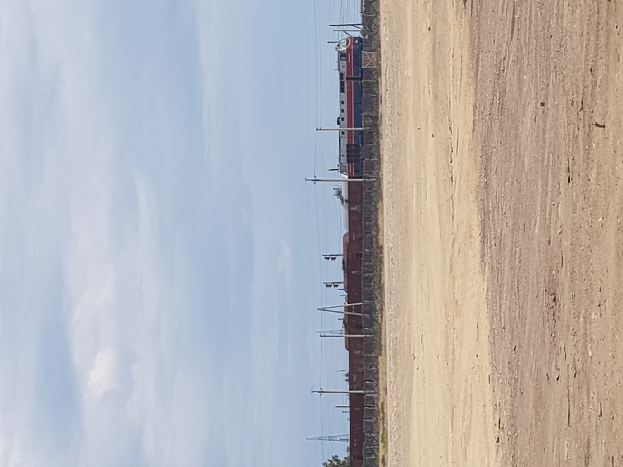 The image shows a train on a railway line in Kurchatov, Kazakhstan. The train appears to be composed of several cars, with a locomotive at the front. The locomotive is painted blue and red, and the cars are red. The train is on a track that runs through a desert-like environment. The ground is sandy and dry, with sparse vegetation. In the background, there are power lines and utility poles. The sky is cloudy and overcast. The scene appears to be industrial or utilitarian, possibly near a railway yard or industrial facility. There are no people visible in the image.