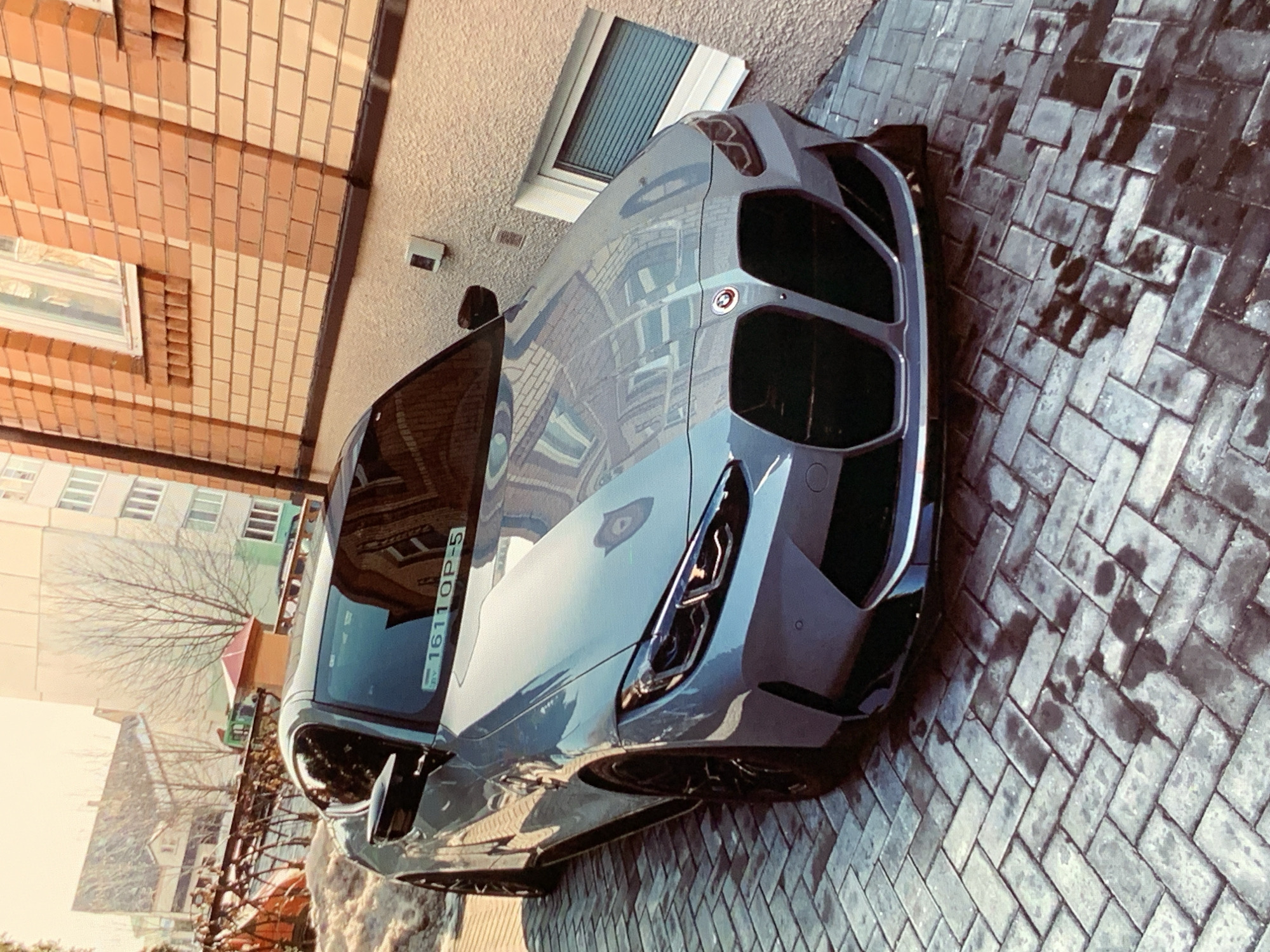 A light blue BMW M4 is parked on a wet cobblestone driveway in front of a brick building. The car is angled towards the camera, with its front end prominently displayed. The building is constructed of orange and light tan bricks, with a white-framed window visible on the right side. In the background, there are apartment buildings and trees. The sky is a bright, clear blue. The license plate of the car is visible and reads "1611OP-5". The cobblestones show dark, wet patches, indicating recent rain or washing.