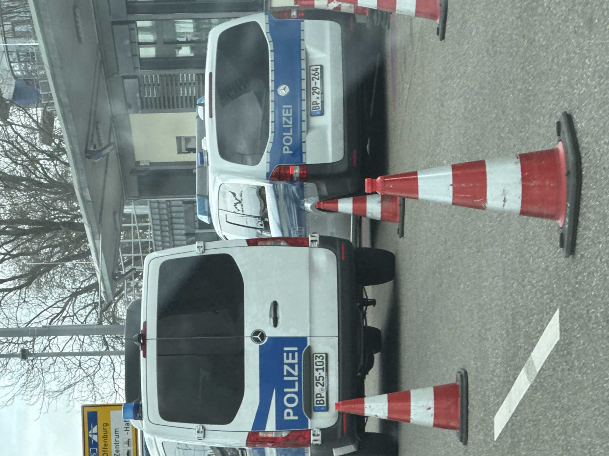 The image shows two white Mercedes-Benz police vans parked on an asphalt surface delineated by red and white striped traffic cones. The vans are marked with "POLIZEI" in blue. The license plate of the front van reads "BP 25-103" and the license plate of the rear van reads "BP 29-264". A sign indicates the location as Offenburg, Zentrum (center) in Kehl, Germany. Bare trees are visible in the background. A building is partially visible behind the vans.