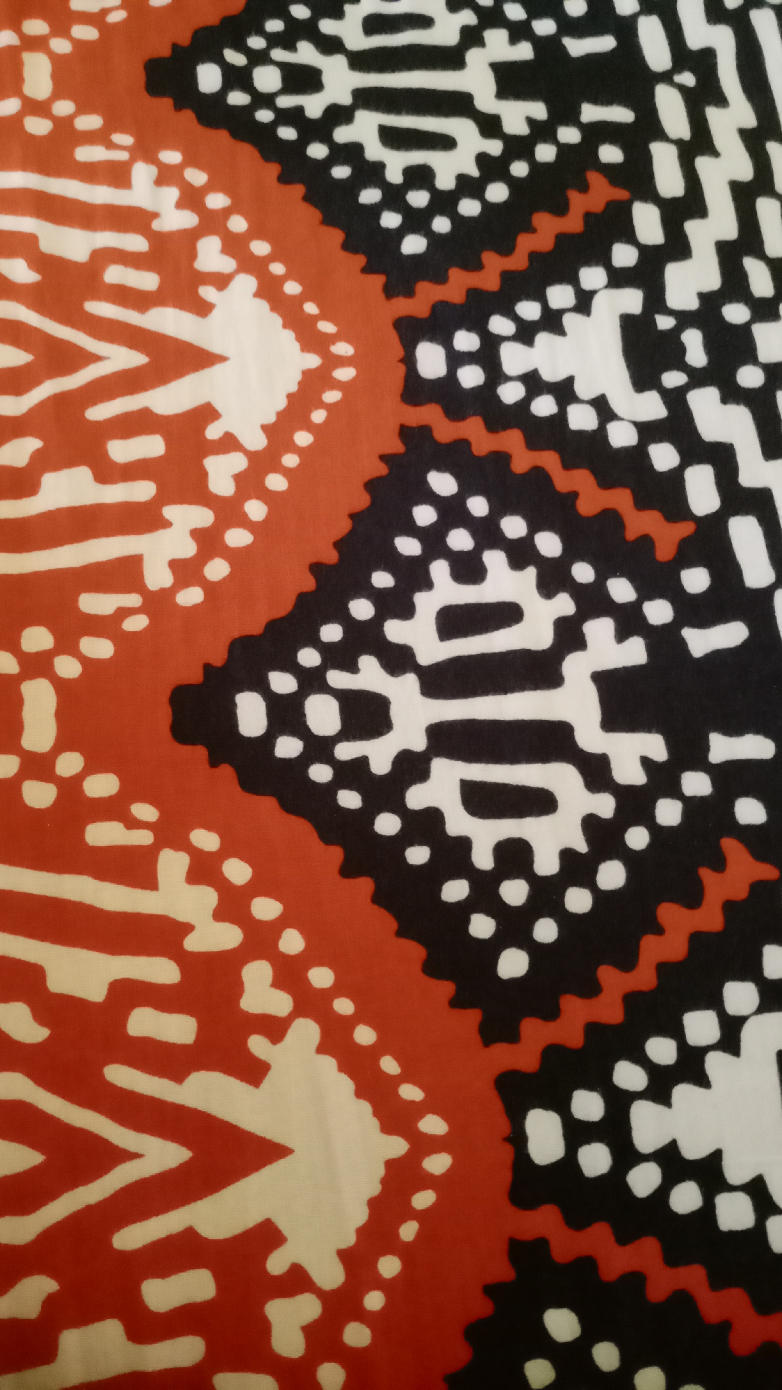 The image displays a close-up view of fabric with a tribal pattern. The pattern features geometric shapes in orange, black, and white. The orange areas have a textured appearance and form large chevron-like designs. These are bordered by a jagged line of black, which then gives way to intricate white shapes surrounded by small black dots. The overall impression is of a vibrant and detailed textile. No specific location within Mertan, Indonesia, can be identified from this image alone.