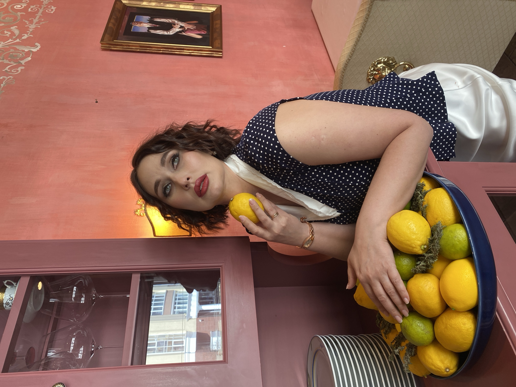 A young woman with dark, wavy hair, light eyes, and red lipstick gazes directly at the camera in this portrait-oriented image. She wears a dark blue polka-dot dress with a white collar or scarf detail. Her left hand holds a single yellow lemon, while her right hand rests within a large blue bowl brimming with numerous yellow lemons, a few green limes, and scattered green foliage.
The setting is an indoor space, characterized by a textured, dusky pinkish-red wall, partially adorned with a silver scrollwork pattern. A gold-framed painting of abstract figures hangs on the wall. To the right, a golden decorative element, possibly from furniture, is visible alongside a white satin-like fabric. Below the woman, a matching pinkish-red cabinet with glass doors displays glassware, including wine glasses. The reflection in the glass reveals parts of urban buildings with windows. Stacked white plates with black stripes are also present. A yellow-orange wall sconce emits a warm glow behind the woman's head. The overall scene suggests a stylish, possibly retro-inspired interior in Yekaterinburg, Russia. No visible text is present.
