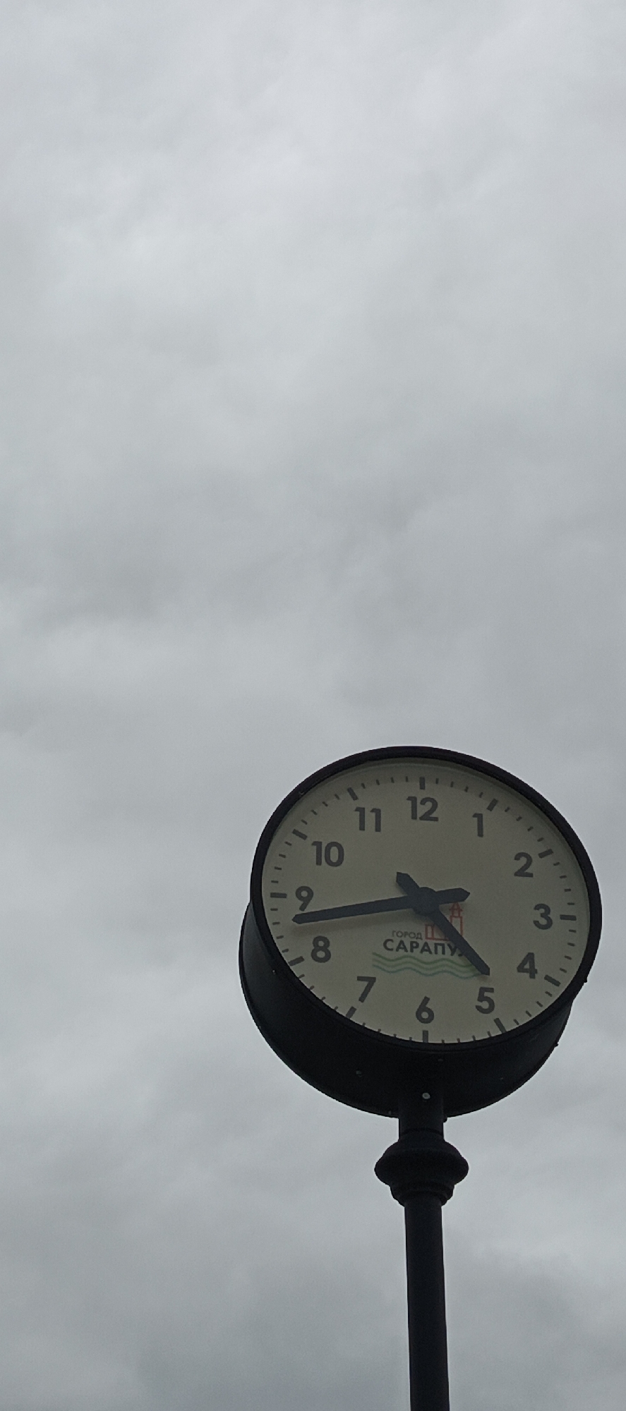 A large, round, dark-framed outdoor clock is prominently featured in a low-angle shot, positioned against a uniformly overcast, gray sky. The clock face is off-white, marked with black Arabic numerals 1 through 12 and minute intervals. The shorter hour hand points just past the 4, and the longer minute hand points to the 8, showing the time as approximately 4:40. The Russian text "ГОРОД САРАПУЛ" (CITY SARAPUL) is printed below the clock's center, identifying the location as Sarapul, Russia. Beneath the text is a logo depicting blue waves with a red and yellow structure. The clock is mounted atop a dark, metal pole that features a decorative collar at its upper section. No individuals or other objects are present in the scene, which is illuminated by diffuse daylight.