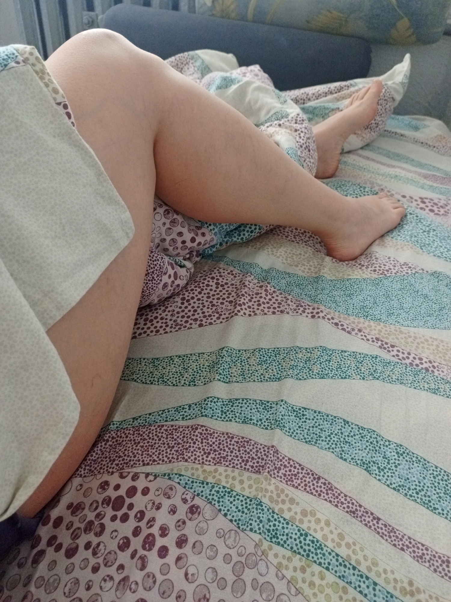 The image displays a close-up view of a person's legs and feet, resting on a bed in an indoor setting, likely in Chaikovsky, Russia. The person appears to be relaxing or lounging.
The foreground is dominated by the person's bare legs, which have a light skin tone. The left leg is bent at the knee, with the thigh partially covered by a light-colored, subtly patterned sheet or blanket. The right leg extends further down the bed, mostly beneath the main bedding, with the foot visible at the bottom right.
The bed is covered with a vibrant, patterned duvet or sheet. It features wide horizontal stripes in varying colors, including light blue, green, purple, light yellow, and white or cream, each adorned with a fine dotted texture. On the lower left part of the bed, the pattern transitions to a design with large, overlapping circles in shades of purple and brown.
In the background, parts of a dark blue or grey upholstered headboard or sofa are visible, along with a patterned cushion. A white, ribbed radiator is partially seen in the upper left corner of the image, suggesting a home environment. The lighting is soft and natural, indicating it is daytime, possibly morning or early afternoon. No text or specific emotions are discernible, but the scene conveys a sense of comfort and tranquility.