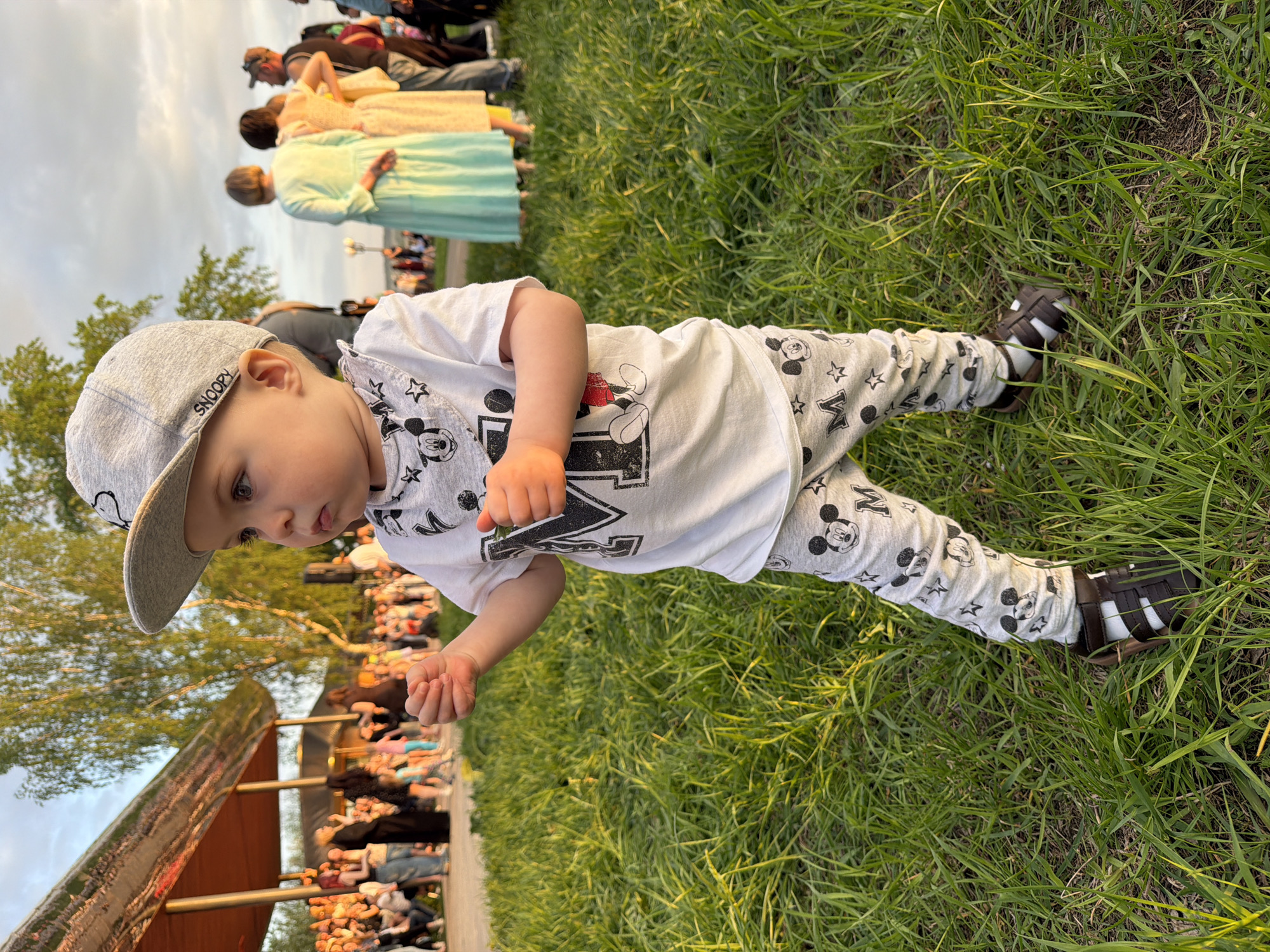 A toddler wearing a backwards gray Snoopy hat, a white Mickey Mouse T-shirt, gray Mickey Mouse pants, and brown sandals sits on green grass. An audience is seated behind a structure. People are standing in the background. The scene is in Votkinsk, Russia.