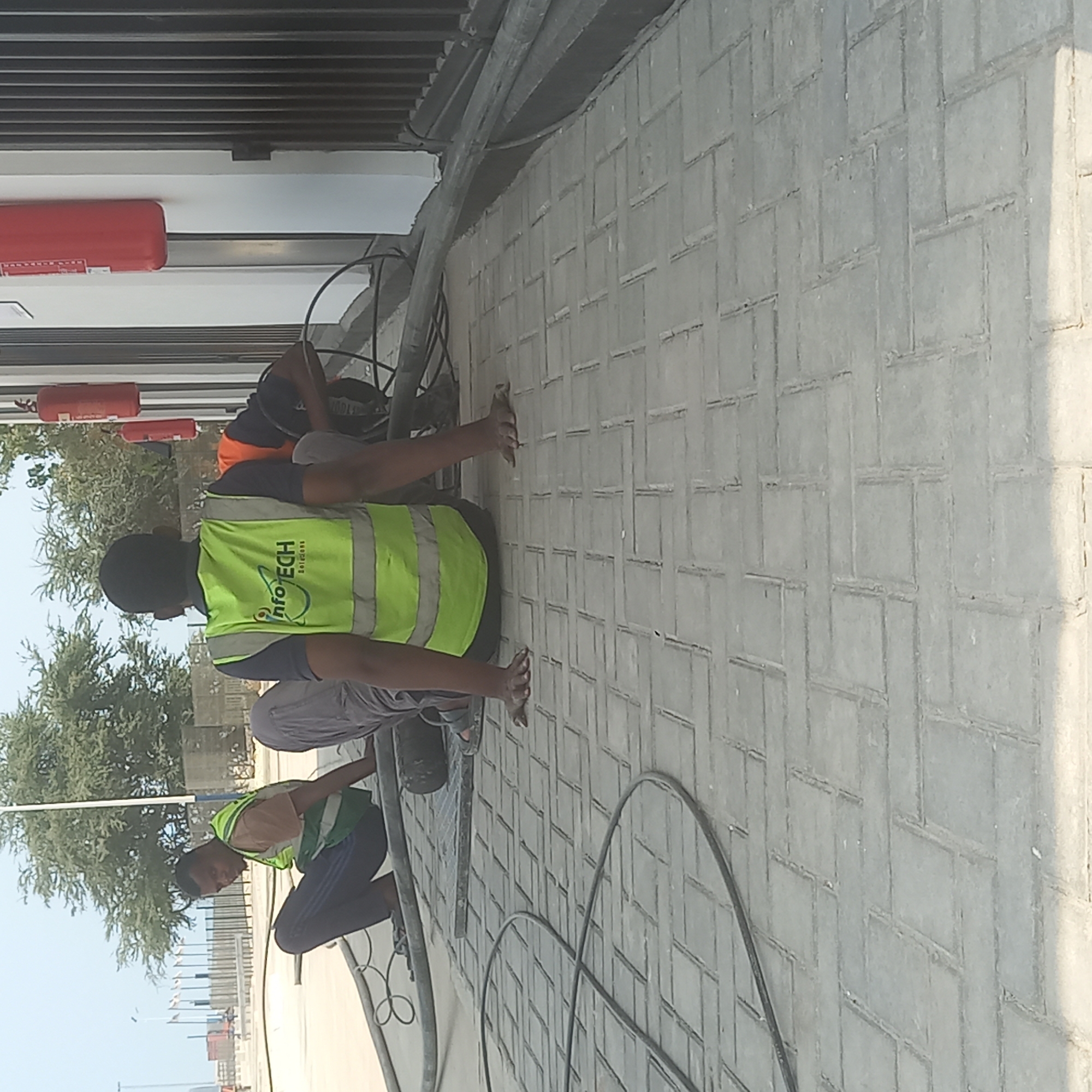 The image captures a scene in Bububu, Tanzania, where three men in work attire are situated on a paved outdoor area. The man on the left, wearing a tan shirt and a green high-visibility vest, is sitting with his legs extended. In the center, another man, also in a green high-visibility vest with the "INFO TECH Solutions" logo visible, is seated with his arms resting on the ground. To the right, a third man, wearing an orange shirt, is partially visible, also seated. All three individuals appear to be engaged in some form of work, with cables and other equipment present on the ground around them. The environment suggests a construction or maintenance site, with paved ground, trees in the background, and what appears to be a building with fire extinguishers mounted on its exterior wall. The overall atmosphere is one of industrial activity.