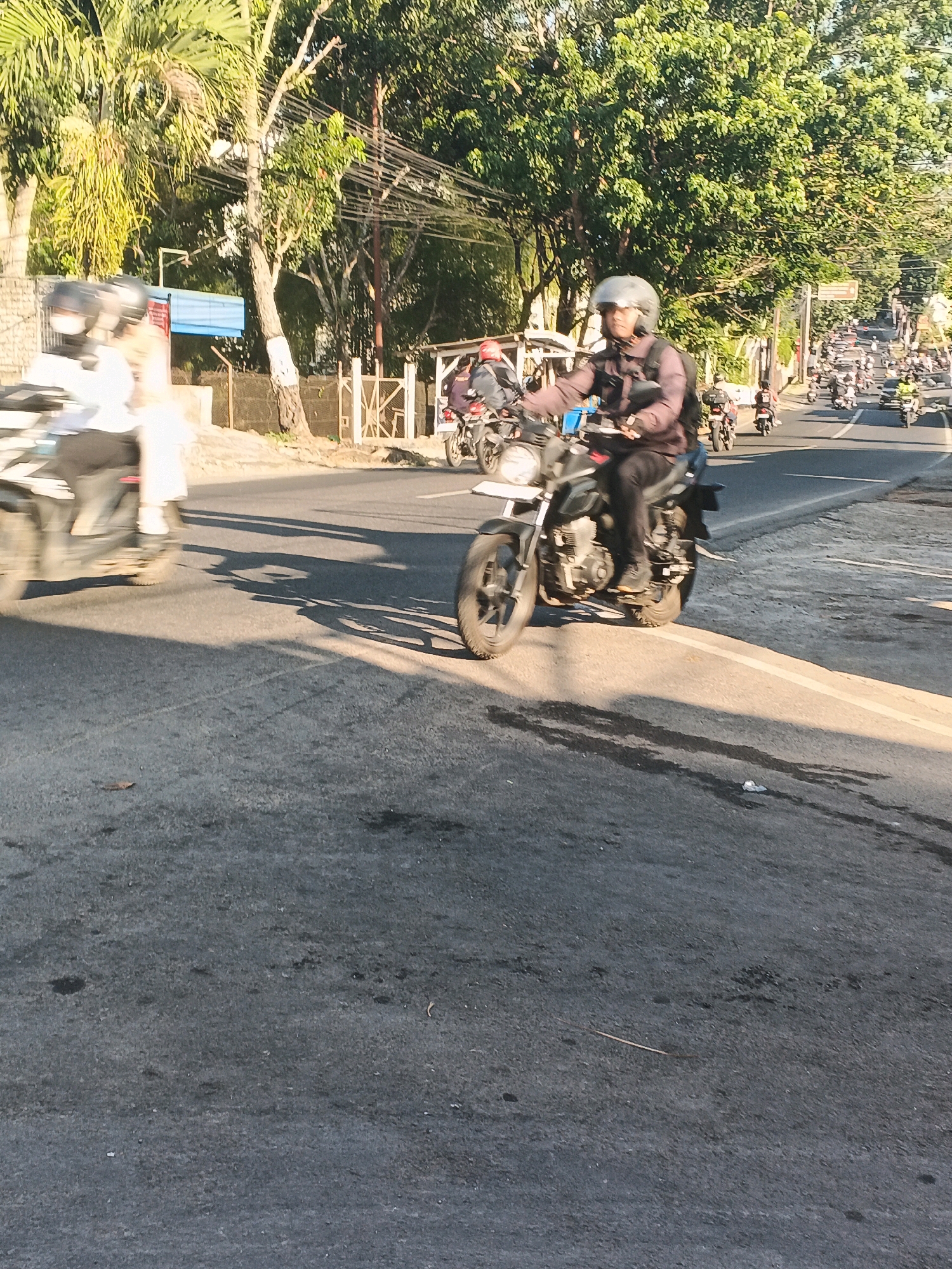 A person wearing a helmet and a dark jacket rides a motorcycle on a paved road. The motorcycle is black with a white license plate. The rider's legs are visible, wearing black pants and shoes. The road has a white line marking on the right side. The background shows trees and a street with other vehicles and pedestrians. The scene appears to be in an urban area in Semarang, Indonesia, with lush green trees and a warm, sunny atmosphere.