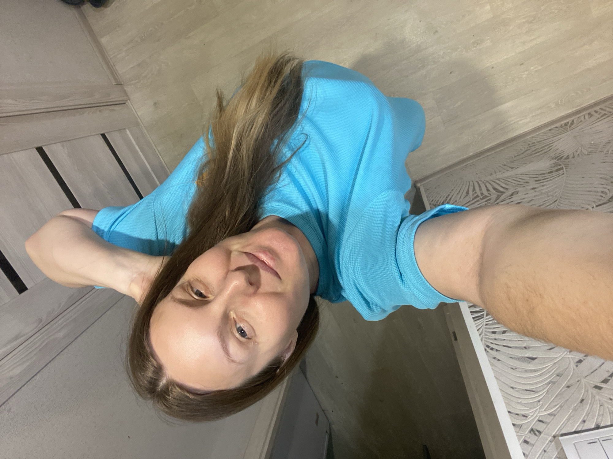 A woman with long, light brown hair is taking a selfie indoors. She is wearing a light blue polo shirt. Her left arm is bent and raised behind her head, while her right arm is outstretched holding the phone. She appears to be in a room with wood-look flooring and patterned wallpaper. The location is in Noviy, Russia.