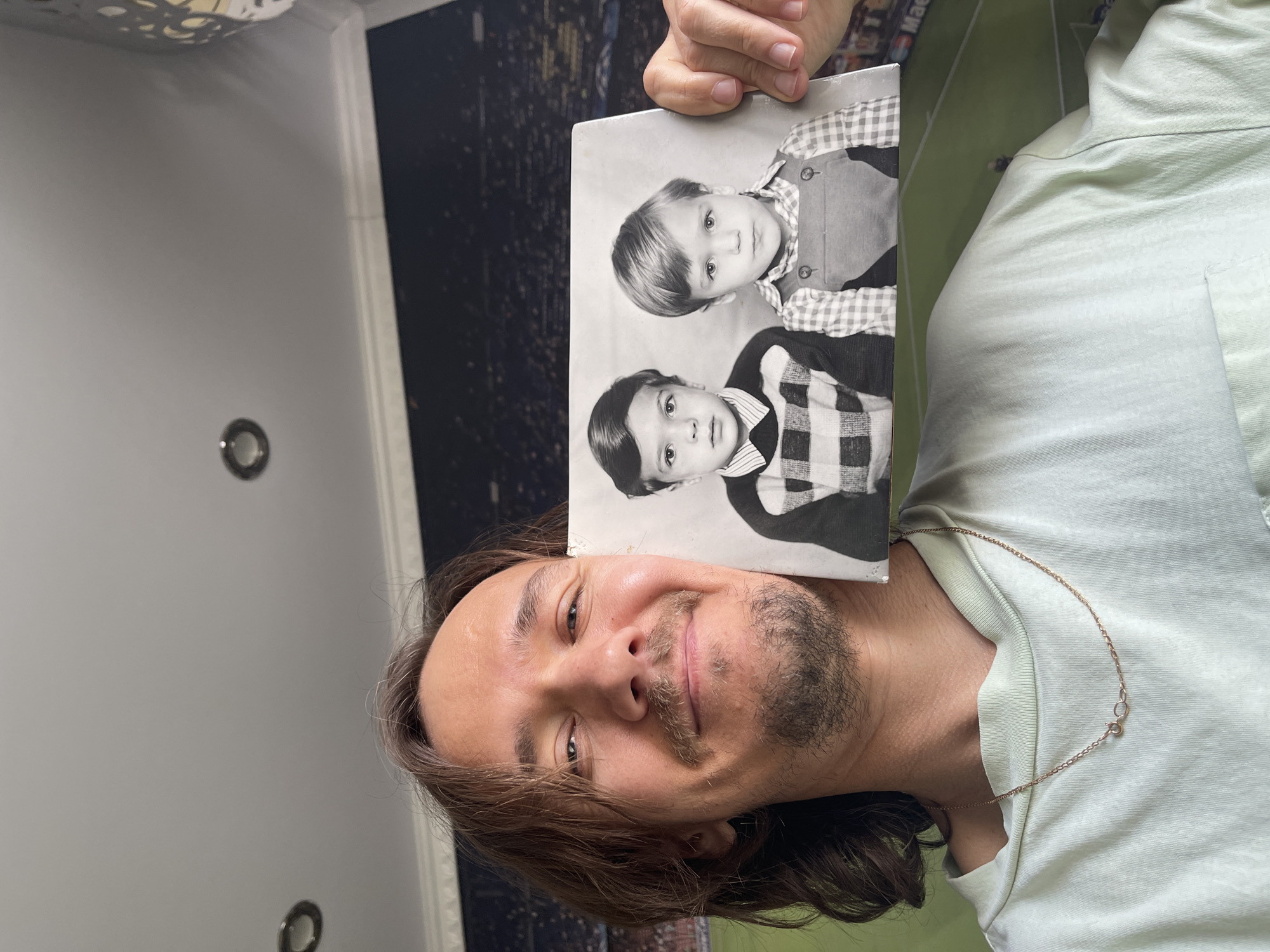 A man with long brown hair, a mustache, and a short beard smiles at the camera while holding up a vintage black and white photograph. He is wearing a light-colored (possibly light green or beige) short-sleeved shirt and a thin chain necklace. His left hand is visible, holding the top edge of the photo.
The black and white photograph features two young boys. The boy positioned higher in the photo has lighter, neatly combed hair and is wearing a plaid shirt with a dark vest. The boy below him has darker hair, parted on the side, and is wearing a checked shirt underneath a patterned (possibly plaid) sweater or jacket. Both boys are looking directly at the camera with serious expressions.
The man's head and the photo are against a varied indoor background. To the left side of the frame, behind his head, is a dark wall with a subtle, possibly city-themed pattern. On the right side, a brighter area reveals a green, patterned wall that resembles a miniature football or soccer field. Two circular recessed lights are visible on a light-colored ceiling. Faint text, possibly "Ma," can be seen on the dark patterned wall behind the man's head, but it is largely obscured. The man's smiling expression suggests a positive or nostalgic mood.