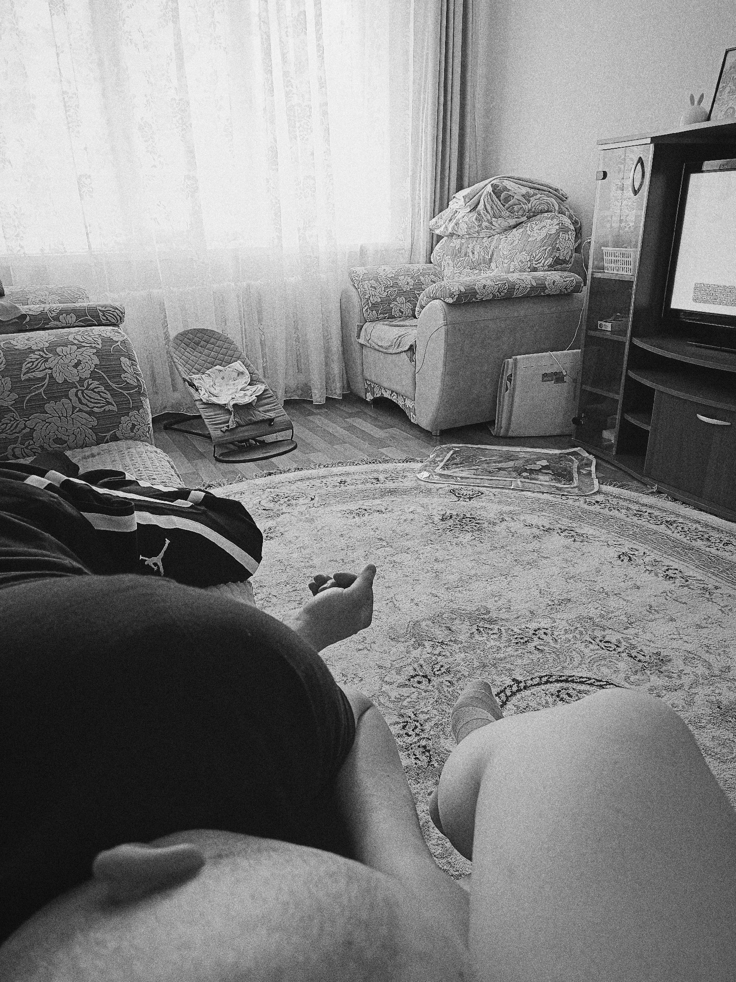 A grayscale, low-angle, interior shot shows a person lying on a patterned rug. The person is wearing dark clothing, possibly shorts or pants, and a light-colored leg is visible. In the background, there is a floral couch, a baby bouncer, a patterned armchair covered with a patterned blanket, a television stand, and a television. Curtains cover the window. The location is likely in a domestic setting. The city of Tobolsk and Russia are not explicitly identifiable.