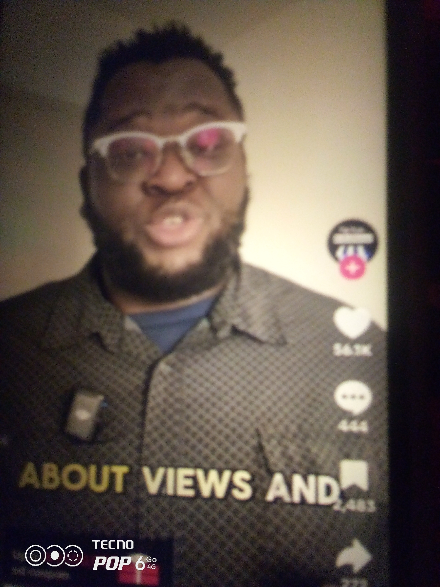 A close-up, slightly blurry image shows a Black man wearing glasses and a patterned shirt. He appears to be speaking. To his right, there are several icons, including a heart with "54.1K" below it, a speech bubble with "444" below it, and a bookmark icon with "2,485" below it. Text in the lower portion of the image reads "ABOUT VIEWS AND" and also displays the "TECNO POP 6 Go 4G" logo. The background is a plain, light-colored wall. The image is of insufficient quality and context to determine its location in Howayi, Nigeria.