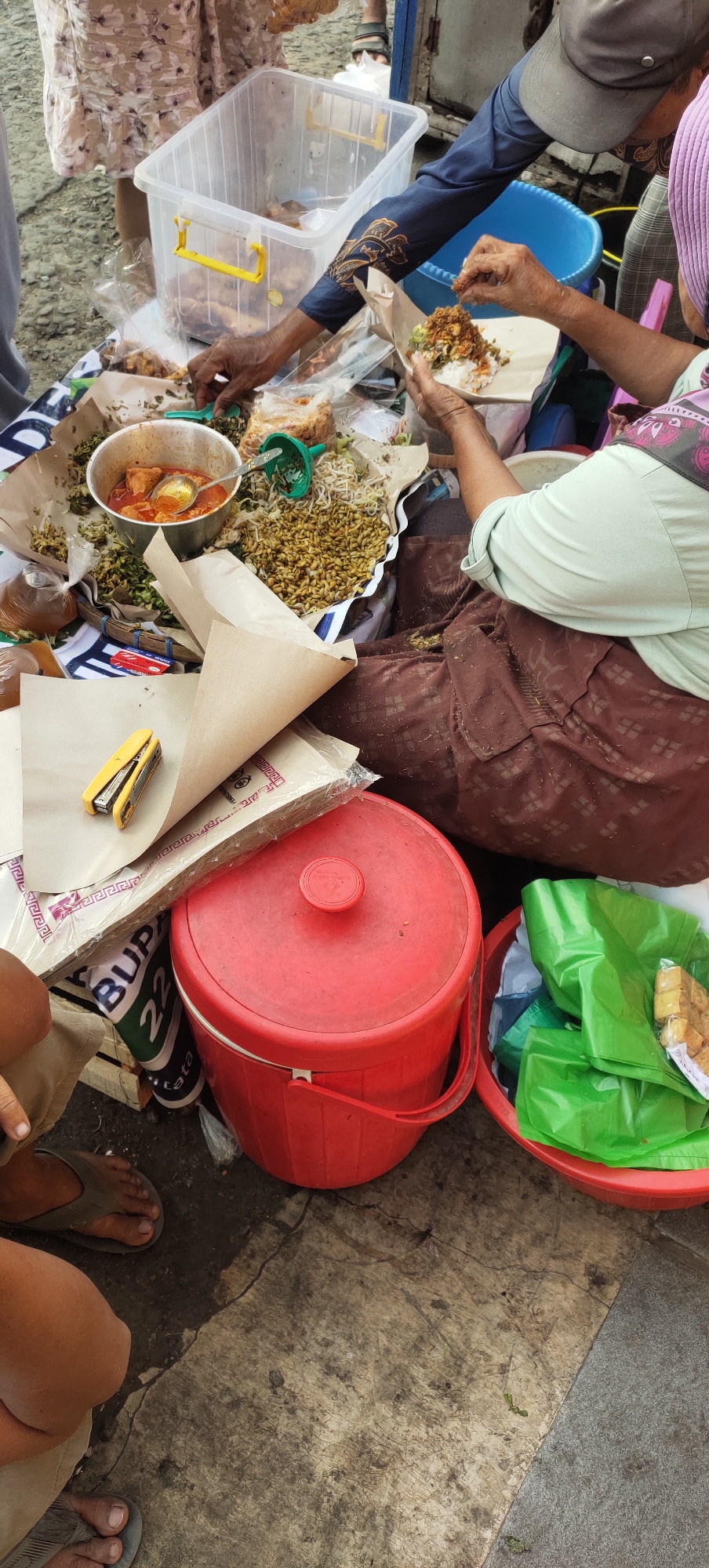 The image depicts an outdoor food stall scene in Karangtowo, Indonesia. In the foreground, a red plastic bucket with a lid is visible, next to a red basin filled with green plastic bags and what appear to be fried snacks. To the left of the bucket, a stack of paper wrappers and a yellow stapler are placed on top of a patterned cloth. Further back, a transparent plastic container with yellow handles holds food items. Several individuals are present, with hands actively engaged in serving or preparing food. One person, wearing a light green long-sleeved shirt and a brown patterned skirt, is being served food on a paper plate. Another person, in a blue long-sleeved shirt with an orange and blue pattern, is reaching towards the food container. The ground appears to be concrete with some cracks and dirt. The overall environment suggests a bustling market or street food setting.
