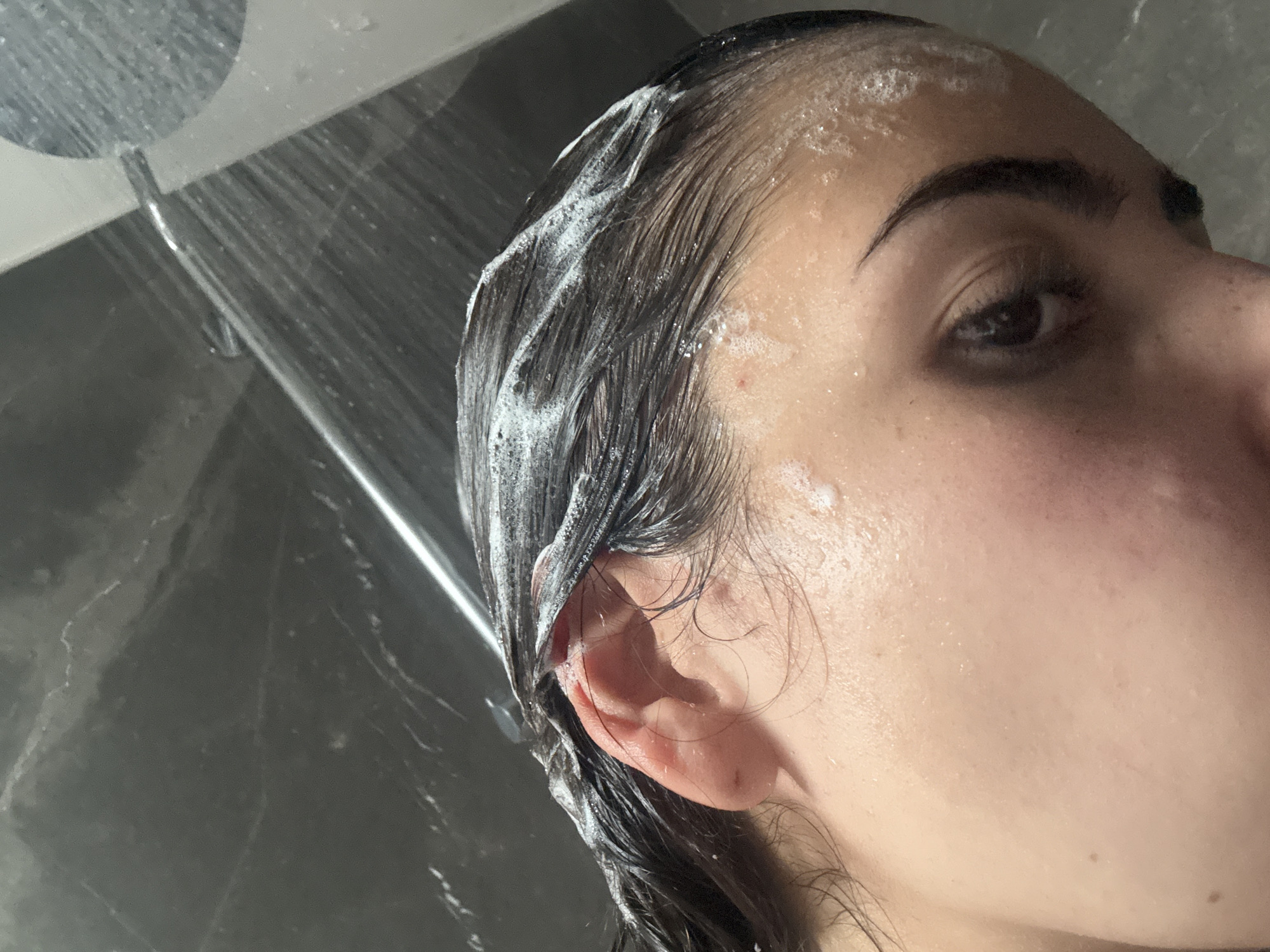 A person with wet, dark hair covered in white suds is in the shower. Water is spraying from the showerhead. Only the side of the person's face is visible. The location is unknown, but the query specified San Bernardino Tlaxcalancingo, Mexico. Therefore, the assumption is that this image was taken in a shower in San Bernardino Tlaxcalancingo, Mexico.