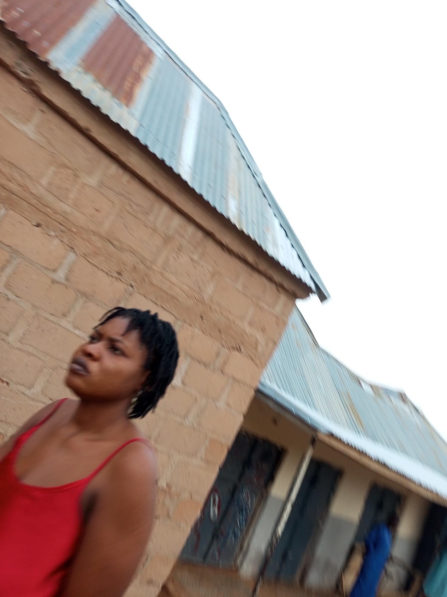 A medium shot captures a young woman with dark, braided hair, wearing a red tank top, looking to her left against a brick wall. The building's corrugated metal roof, showing signs of rust, extends into the frame. To the right, a low-slung building with multiple doorways is visible, with a person in a blue garment partially obscured in one doorway. The overall setting appears to be outdoors in a residential or commercial area in Tempure, Nigeria.