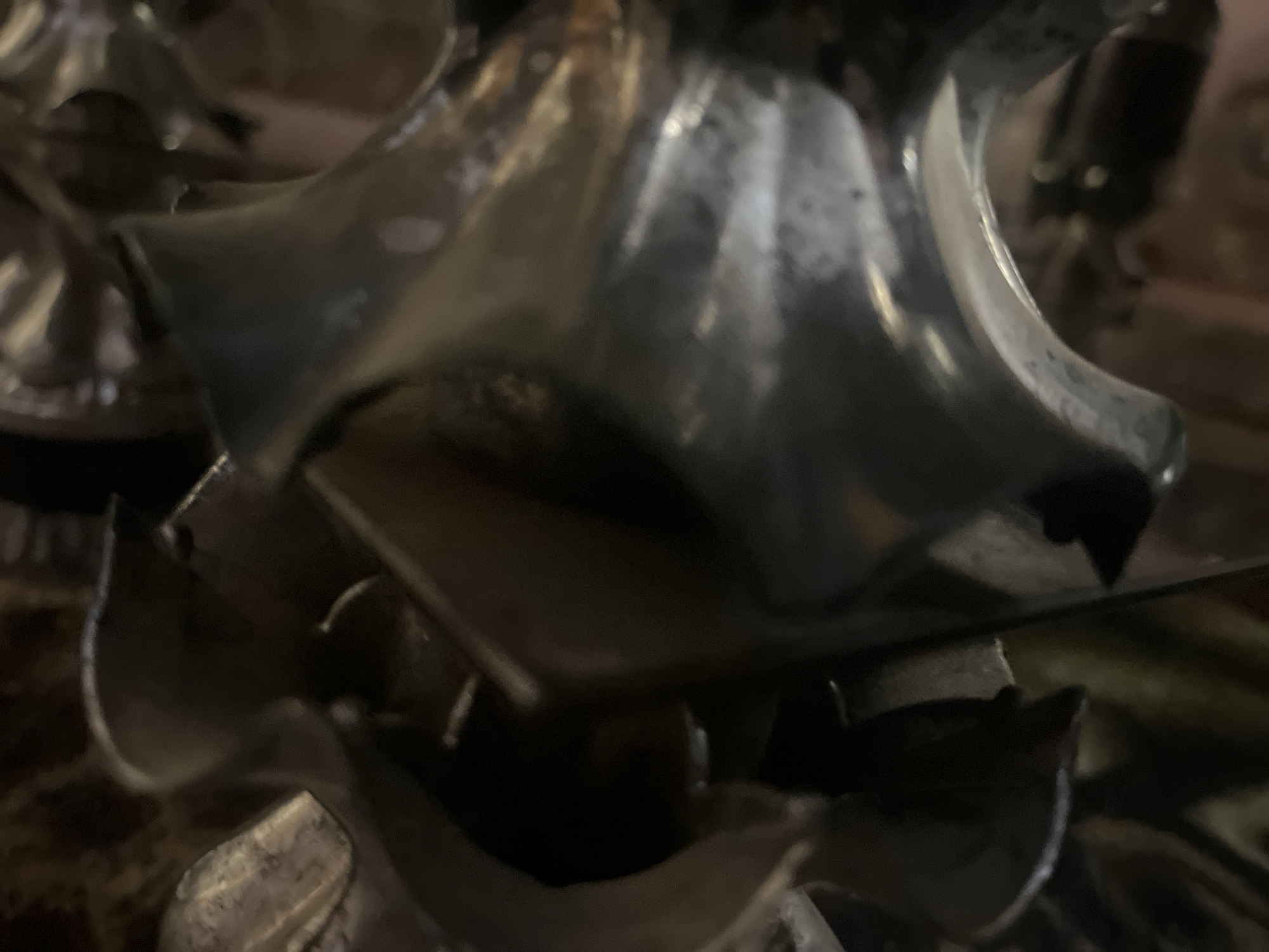 The image shows a close-up, blurred view of metallic objects, possibly parts of a decorative fixture or sculpture, with a dark, textured background. The lighting creates reflections on the metallic surfaces. There is no discernible information about the location in Jalingo, Nigeria, or any people, actions, or interactions. The scene is abstract and lacks contextual detail.