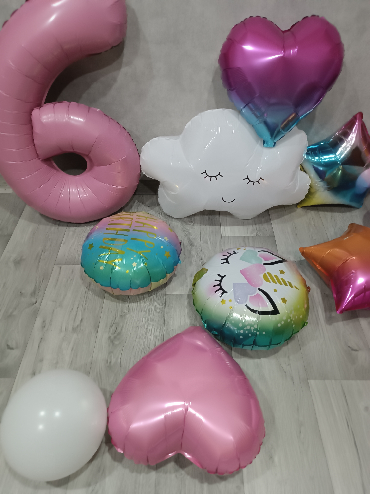 The scene, located indoors in Unknown, Unknown, displays an assortment of inflated balloons resting on a light grey wooden floor against a textured grey wall.
On the left, a large, pink foil balloon shaped as the number "6" leans against the wall. To its right, partially elevated against the wall, is an ombre pink-to-blue heart-shaped foil balloon. Below this heart, a white cloud-shaped foil balloon with closed eyes and a smiling mouth is positioned. Further right, a multi-colored star-shaped foil balloon with a blue-to-yellow gradient is partially visible against the wall.
On the wooden floor, in the foreground, several balloons are distributed. A round foil balloon, displaying "HAPPY BIRTHDAY" text and star motifs on a blue-to-pink gradient background, is centrally located. Adjacent to it lies another round foil balloon featuring a unicorn head design with a green-to-pink gradient. To the mid-right, an orange-to-pink gradient star-shaped foil balloon is present. In the lower left foreground, a plain white spherical latex balloon rests next to a solid pink heart-shaped foil balloon. All balloons appear stationary.