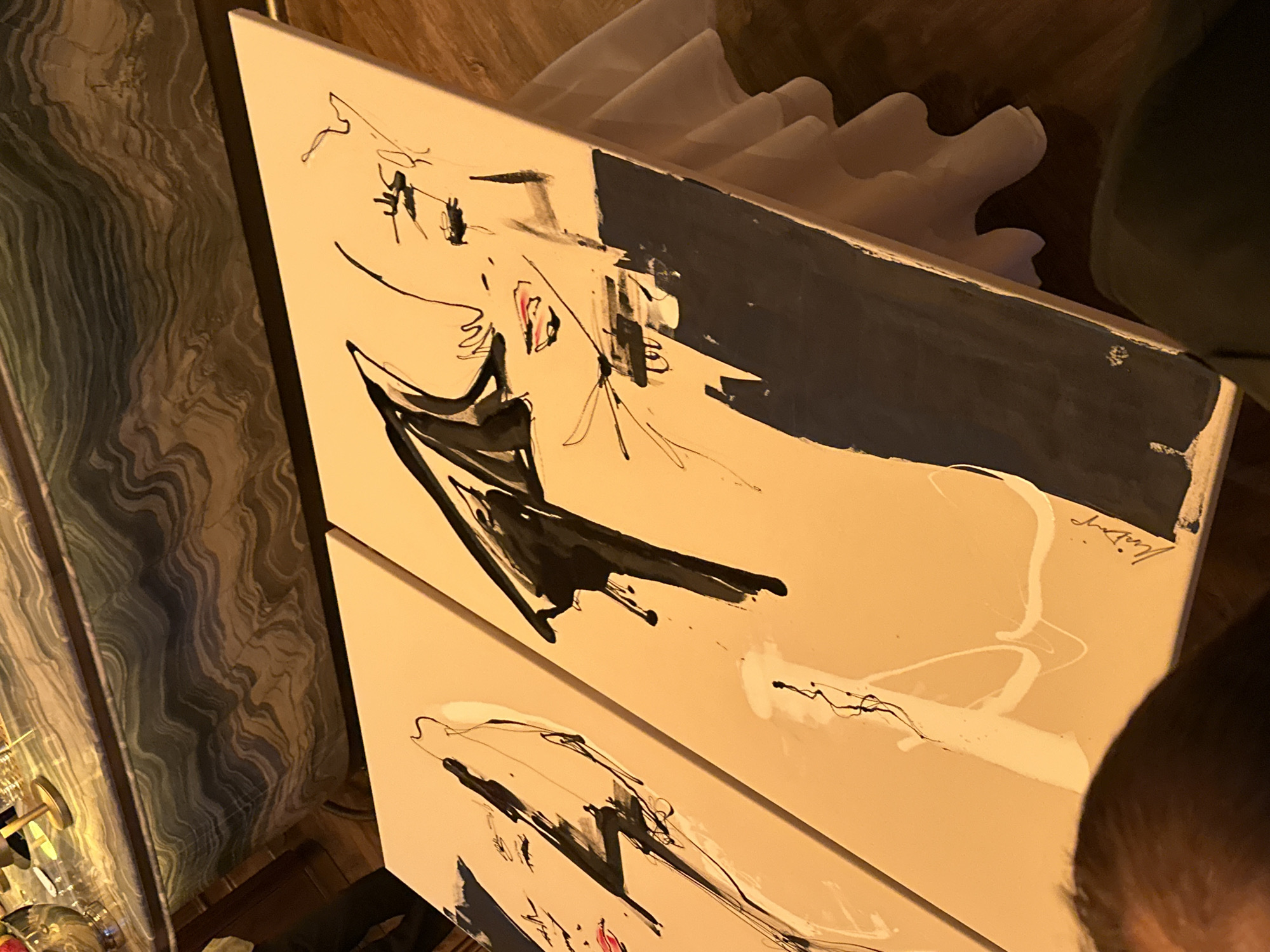 The image shows two abstract paintings displayed on easels or stands, with the top edge of a table visible in the background. The paintings feature fluid lines and bold strokes of black and white paint, with accents of red on the lips of what appear to be stylized faces. The right painting is larger and has a significant portion of dark blue or black paint at the bottom. The background table has a patterned surface with wavy lines in shades of green and gray. The floor is made of wood. The lighting in the scene is warm and appears to be artificial. There is a white cloth draped over the base of the display in the foreground. A person's head and shoulder are visible at the bottom left of the image, and another person is partially visible in the bottom right. The overall scene suggests an indoor exhibition or event space. There is no information within the image to suggest a specific location in London, United Kingdom.
