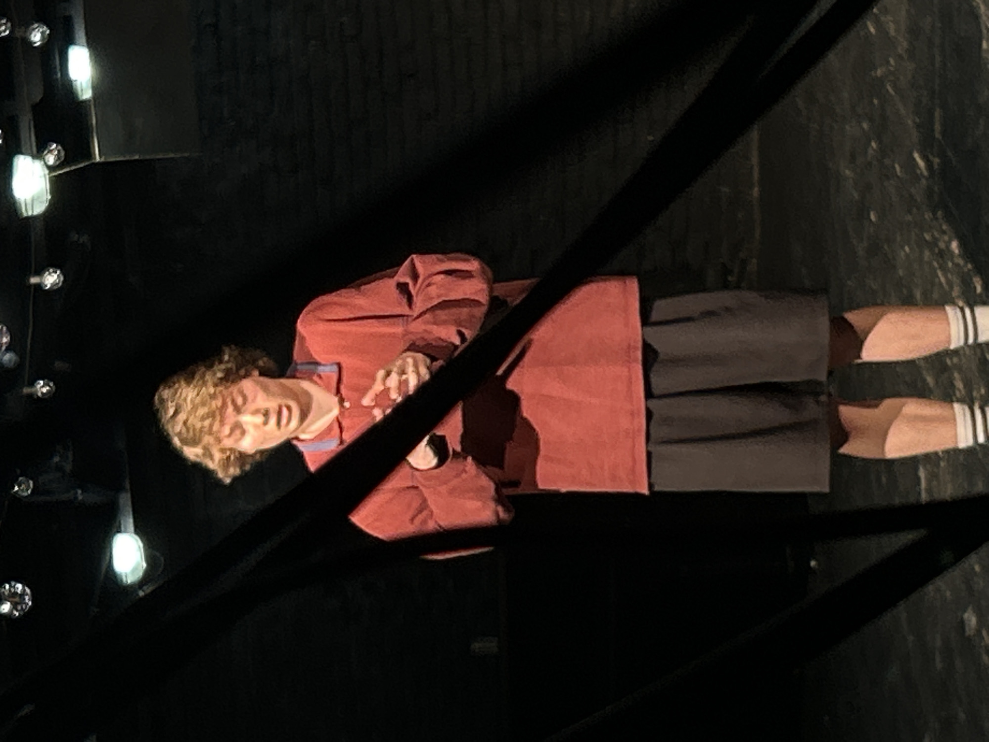An individual is depicted standing indoors, likely on a stage or within a performance setting, with the image oriented vertically. The person has curly, light brown hair and is wearing a reddish-brown long-sleeved shirt with subtle blue striped detailing on the collar or placket. They are also wearing dark shorts or a dark skirt and white socks with black stripes near the top.
The individual is looking slightly downwards and to their left (from the viewer's perspective), with their hands clasped or resting in front of their waist. The lighting is focused on the person, creating a stark contrast with the very dark, almost black background, which appears to be a curtain or wall. On the left side of the frame, several bright, spherical lights with metallic bases are visible, suggesting stage lighting or a backstage vanity mirror setup.
Dark, diagonal elements, possibly stage rigging or architectural features, traverse the foreground, partially obscuring the lower body of the individual. The overall ambiance is that of a dimly lit performance or rehearsal space. No discernible text is visible in the image.