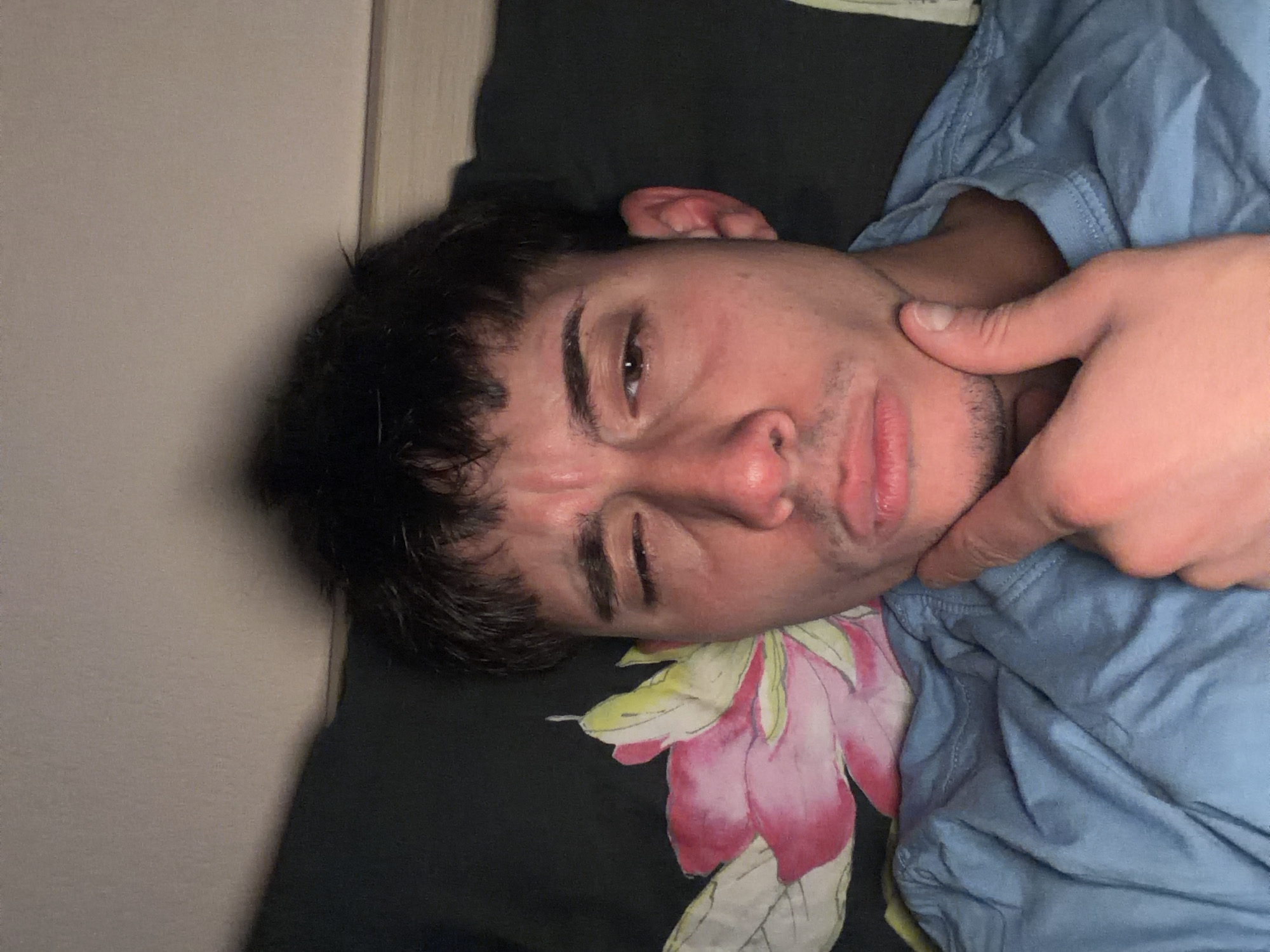 A man is lying on a bed. He has dark hair and is wearing a blue shirt. A hand is touching his chin. The bed has a black pillow with a pink and white flower design. A door is visible to the left. There is no indication in the image that reveals any information about Blagoevgrad, Bulgaria.