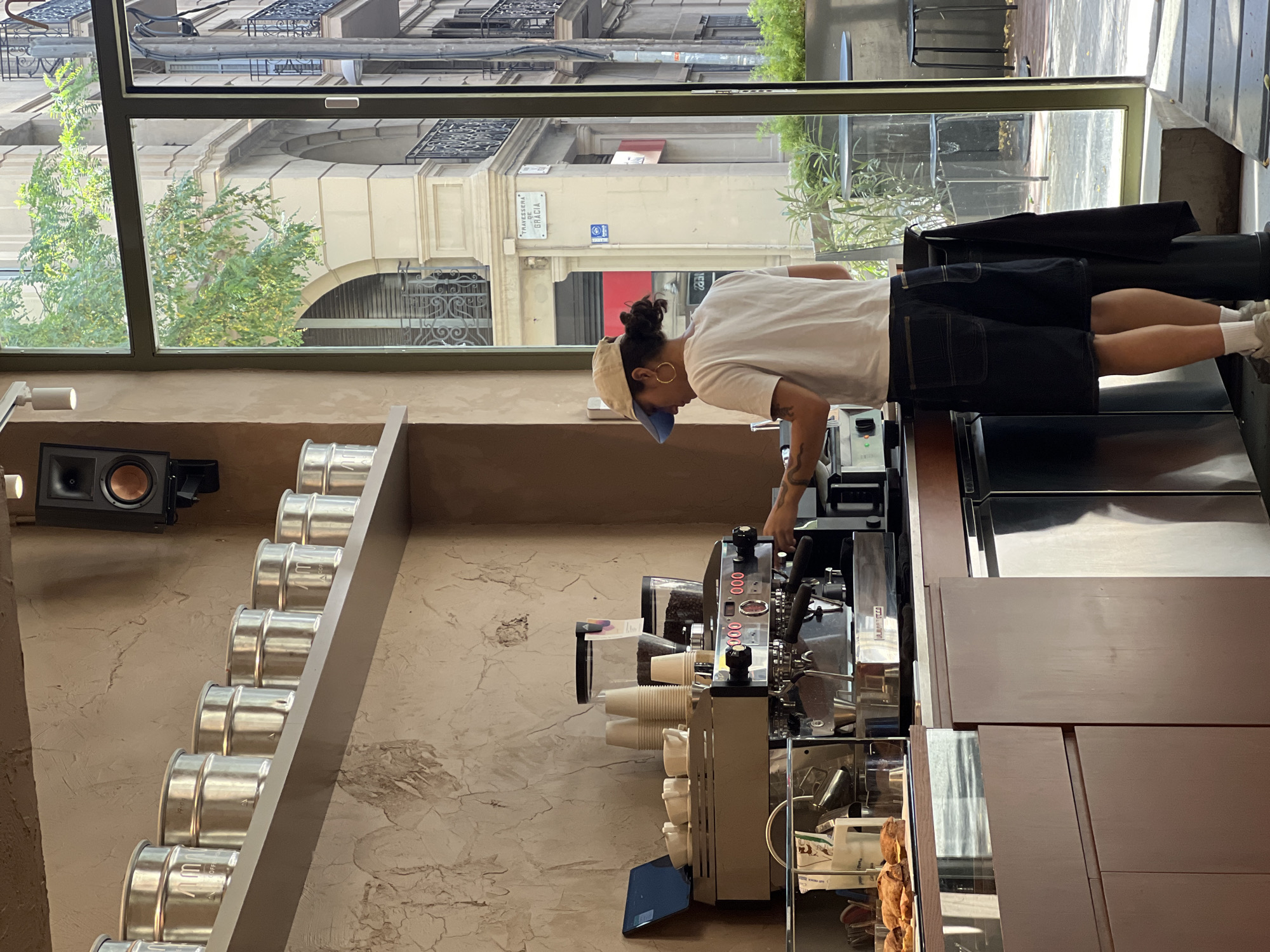 An overhead shot captures a coffee shop in Barcelona, Spain, bathed in natural light filtering through a large window. The scene is dominated by a gleaming espresso machine, where a person wearing a white t-shirt, denim shorts, and a tan baseball cap is actively engaged in preparing a beverage. The person's actions are centered around the machine, with one hand near a portafilter. Several white paper cups are stacked neatly on top of the espresso machine, alongside containers of coffee beans. Above the counter, a row of identical, silver coffee canisters are displayed on a shelf. To the left, a glass display case showcases pastries. The window offers a glimpse of the bustling street outside, featuring architectural elements like arches and wrought-iron balconies, and a street sign that reads "TRAVESSERA DE GRÀCIA." The overall impression is one of a busy, modern coffee shop in an urban setting.
