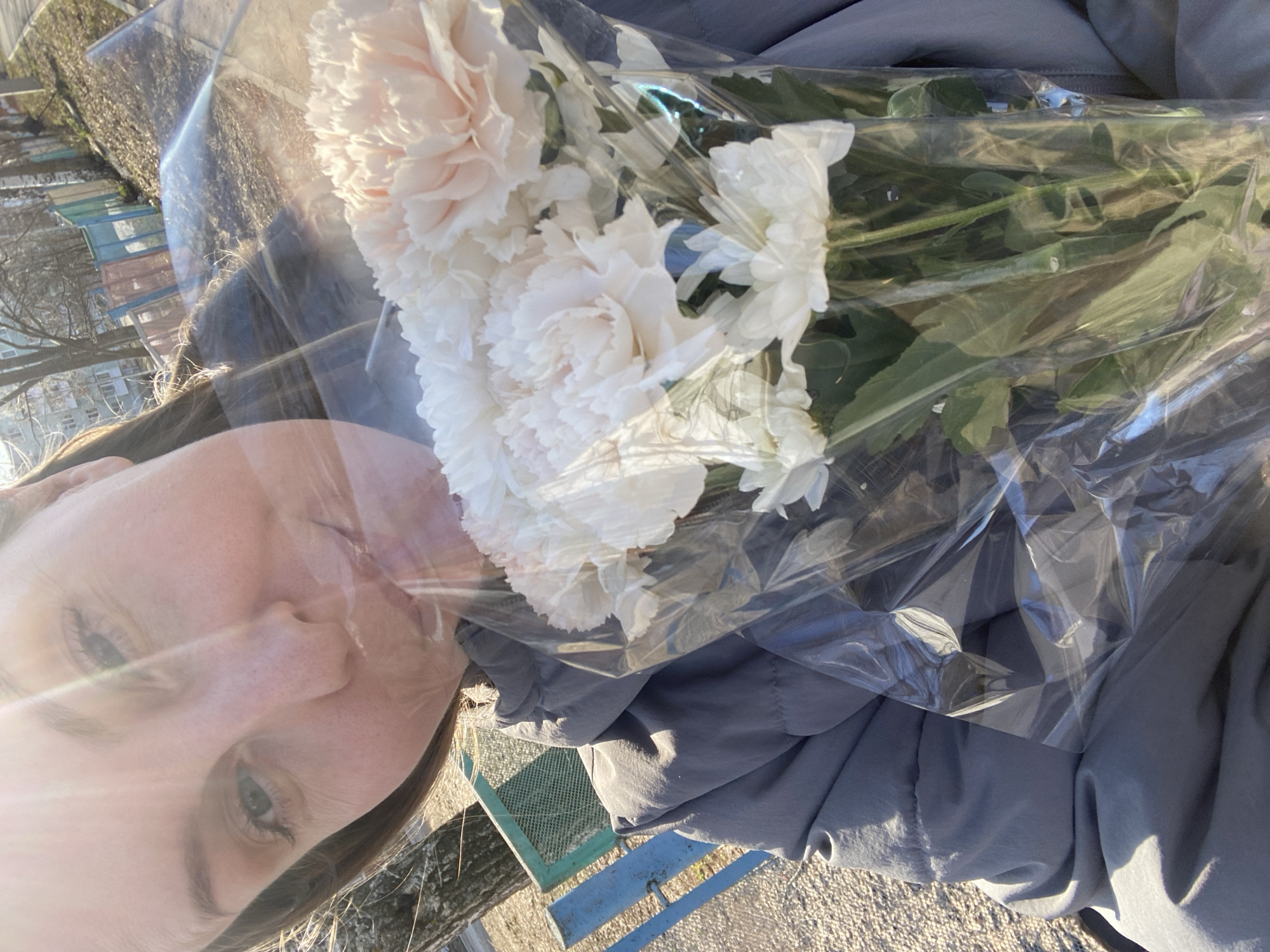 A woman holds a bouquet of white and pale pink carnations wrapped in clear plastic. The woman's face is partially visible, showing her nose, eyes, and forehead. She has fair skin and light hair. The flowers are held against a gray jacket. The background includes a blue and green bench or railing, a glimpse of buildings and trees. The scene appears to be outdoors, possibly in Noviy, Russia.