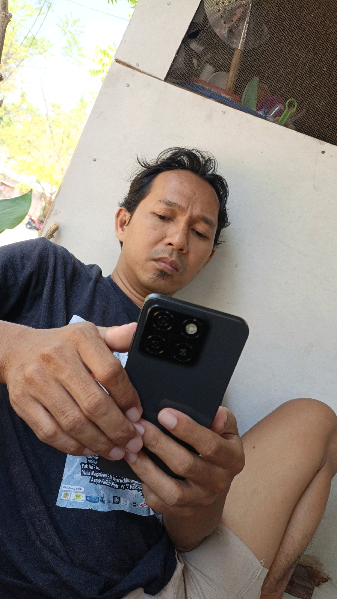 A man is holding a black smartphone with both hands and looking down at it. He has dark hair, a mustache, and a goatee. He is wearing a dark blue t-shirt with a graphic print on the left chest area. He is sitting with his legs crossed, with his right leg extended and his left leg bent. He appears to be outdoors in a semi-rural or residential area of Cikal, Indonesia, with a pale wall and some greenery visible in the background. The lighting suggests it is daytime.