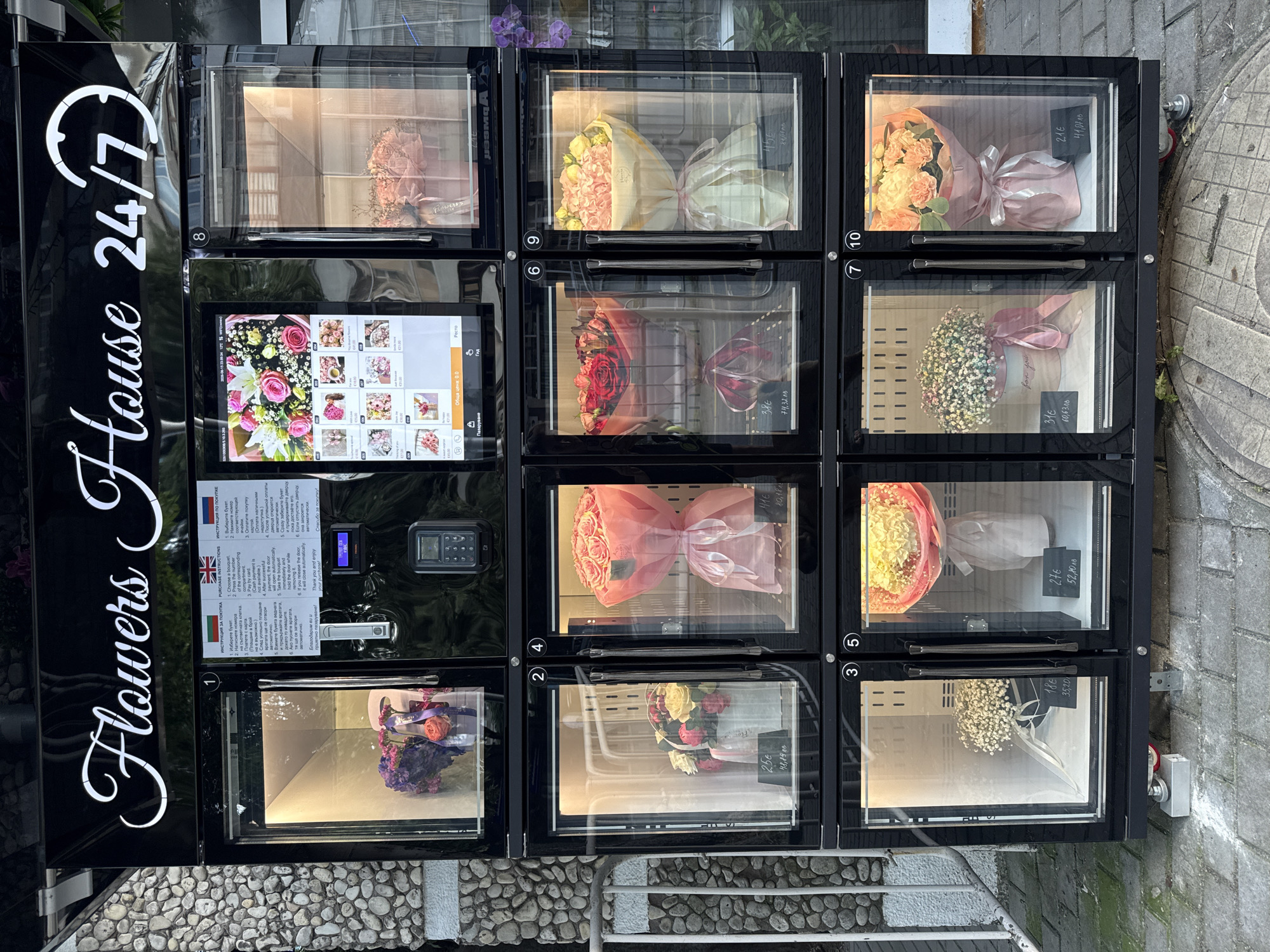 An outdoor flower vending machine, labeled "Flowers House 24/7," is situated on a paved sidewalk in Varna, Bulgaria. The machine is divided into multiple transparent compartments, each displaying a bouquet of flowers. The compartments are numbered, and some have price tags. The machine has a digital screen showing flower options and purchase instructions in multiple languages. The background includes a stone wall on the left and a building on the right. The lighting suggests it is daytime. There are no people visible in the image.