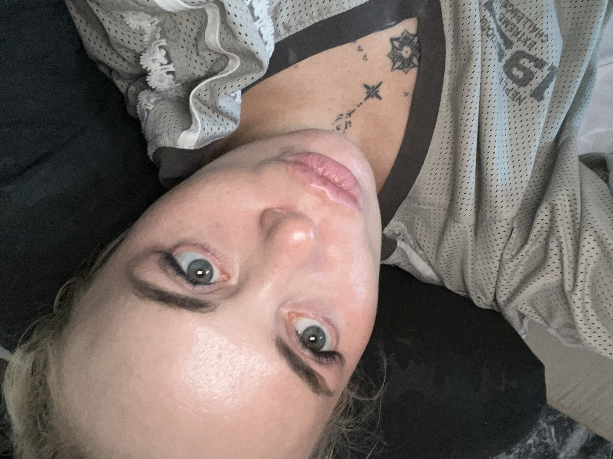 A woman with blonde hair pulled back is shown from a close-up, tilted perspective. She has blue eyes, visible eyebrows, and is wearing makeup, including false eyelashes. Her lips are pink and slightly parted. A tattoo is visible on her chest. She is wearing a grey mesh top with white ruffled trim. The background is dark and indistinct. The scene appears to be indoors.
