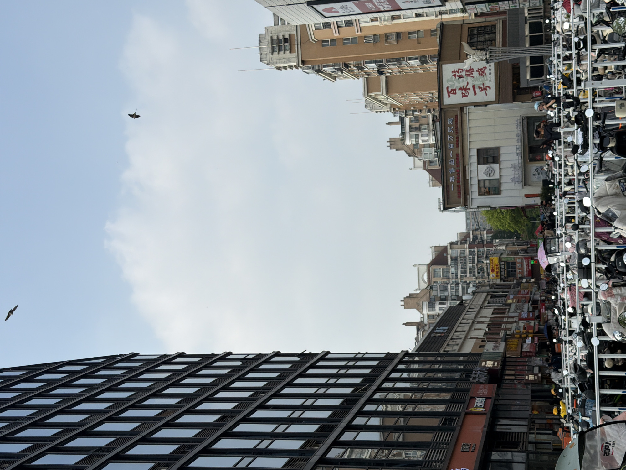The image captures a vibrant urban street scene in an unnamed Chinese city, likely Jiangning District based on context. Although presented vertically, the scene depicts a horizontal street perspective.
In the foreground and to the right, numerous scooters and motorcycles are densely parked in neat rows, with some appearing more haphazardly placed. Many helmets are visible resting on the seats, suggesting busy activity. A metal barrier or rack helps organize the vehicles along what appears to be a sidewalk.
Beyond the parked vehicles, the street is lined with a mix of buildings. On the right, older, multi-story buildings house various businesses, indicated by numerous colorful signs adorned with Chinese characters. Visible text from these signs includes "布丁酒店" (Pudding Hotel), "一年售出一百万只鸡" (One million chickens sold per year), "百味一号" (Taste No. 1), "药膳鸡" (Herbal Chicken), and "二十年老店" (Twenty-year-old store). Further down the street, more buildings and distant cityscapes recede.
On the left side of the scene, a prominent, modern building with a dark, grid-patterned glass facade stands tall, reflecting the bright sky. The sky above is partly cloudy, with patches of bright blue. Two objects are visible in flight: a dark, distinct kite shaped like a manta ray or stingray, and a smaller, dark bird.
The scene depicts a bustling daytime environment. The light is bright but diffused, suggesting either an overcast day or perhaps late morning/afternoon. A few people are faintly visible on the street and near the scooters, including one person wearing a helmet, hinting at the everyday life of the city. The architectural styles, the abundance of scooters, and the Chinese script on the signs are strong cultural and geographical indicators.