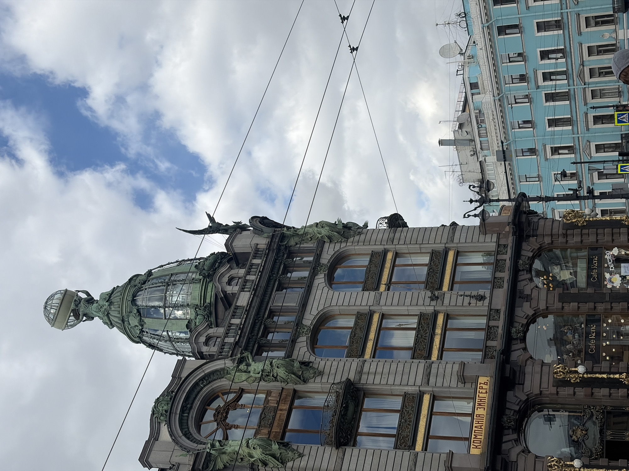 The image depicts the Singer House in Saint Petersburg, Russia, situated against a backdrop of a cloudy sky. The architectural details of the building are prominently displayed, highlighting its ornate dome with a glass and metal framework, topped by a sculpture of a woman holding a globe. Sculptural embellishments adorn the façade, particularly around the arched windows. A sign displaying the name "КОМПАНИЯ ЗИНГЕРЪ" is visible on the building's side. The building's lower levels have signage that reads "Cafe René." A neighboring blue building is also captured in the background. Utility wires are visible, running diagonally across the sky.