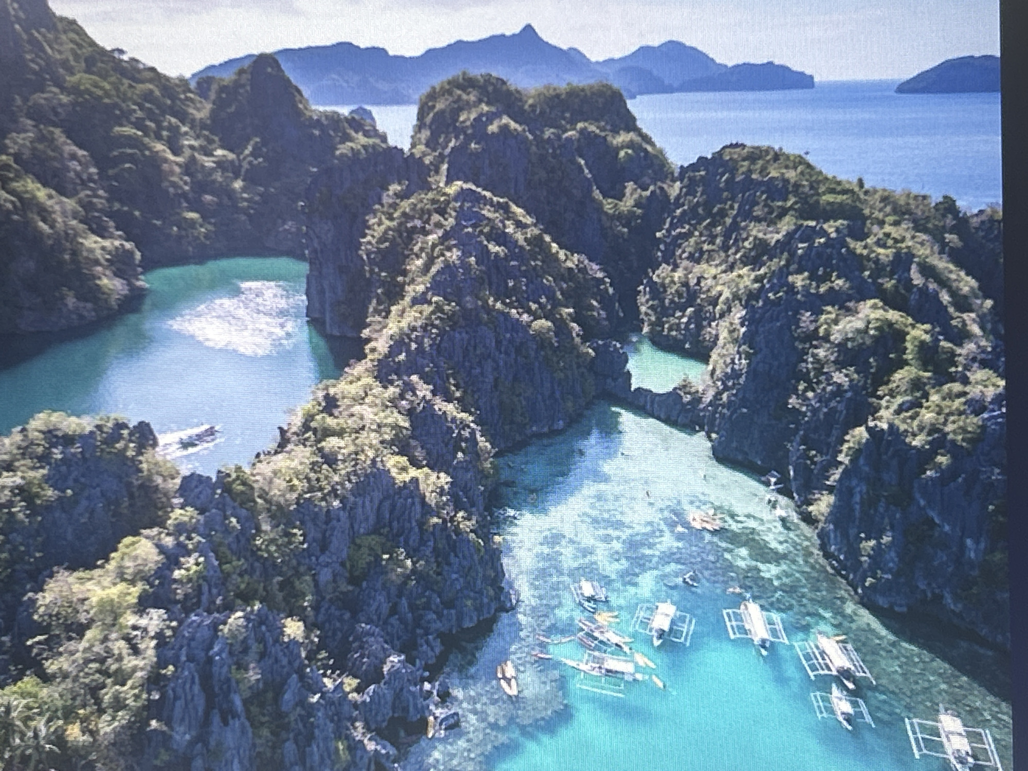 This aerial photograph captures a tropical island landscape. Jagged, dark green, rocky cliffs covered with vegetation rise from vibrant turquoise waters. Several outrigger boats, appearing to be traditional Bangka boats, are scattered across the shallow, clear water, some with people visible. In the distance, more islands and a mountainous horizon are visible under a bright sky. The scene depicts a serene, natural environment, likely a popular tourist destination for water activities and island hopping. The image does not depict Sofia, Bulgaria.