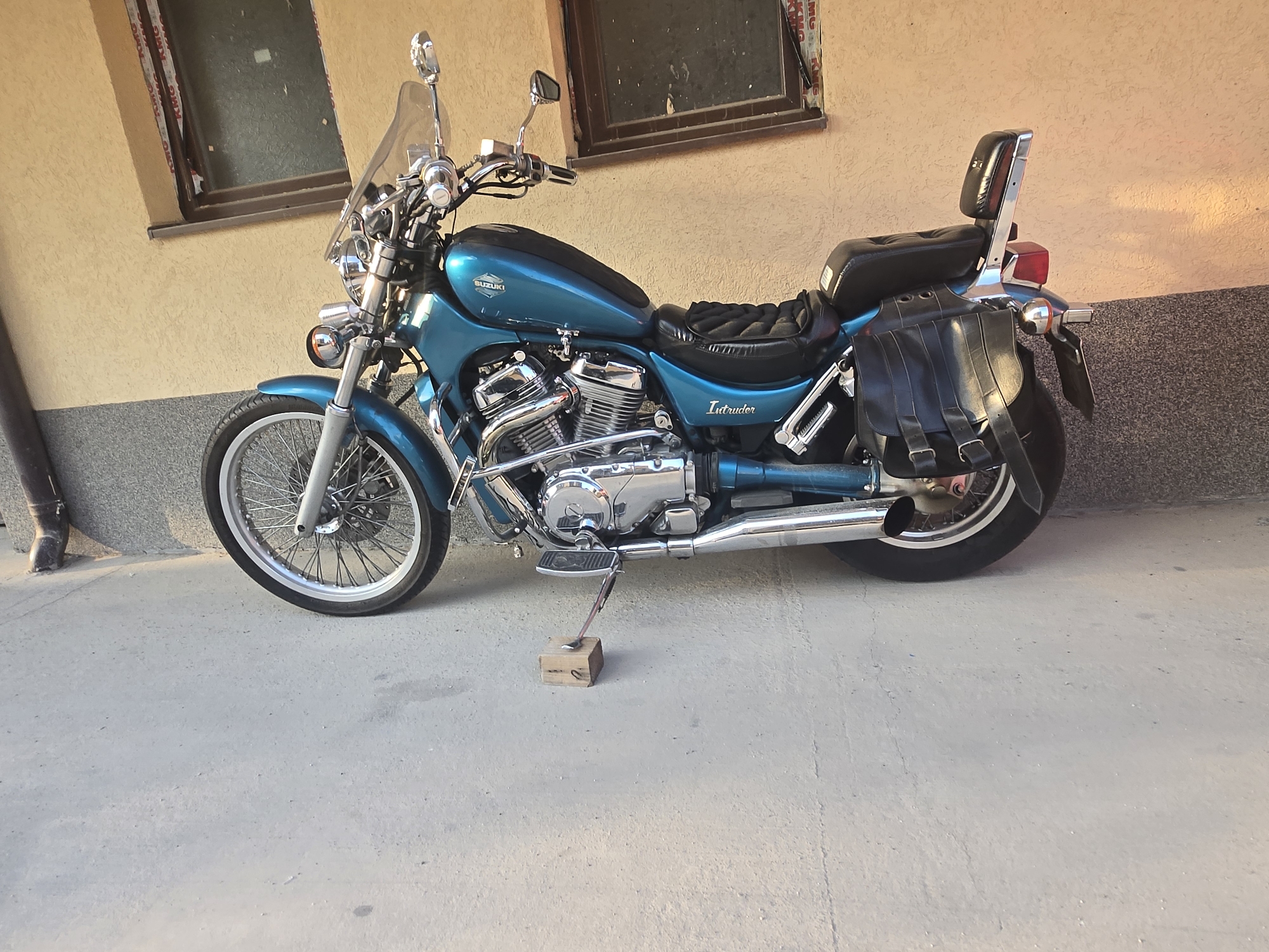 A light blue Suzuki Intruder motorcycle is parked on a concrete surface next to a beige wall with two windows. The motorcycle has a chrome engine, chrome exhaust pipes, and black leather saddlebags. A small wooden block supports the kickstand. The location is Unknown, Unknown.